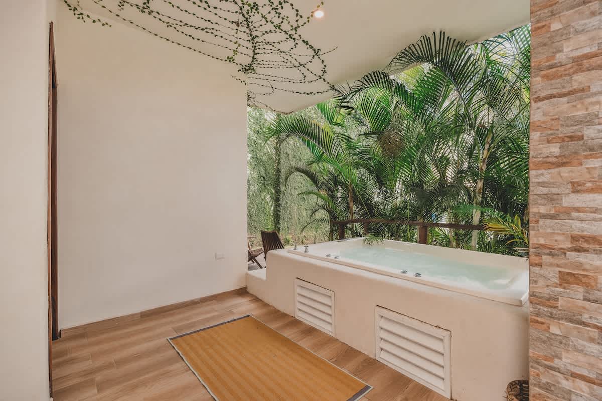 A spacious outdoor patio features a built-in white hot tub, a brown floor mat, and white walls, with lush green tropical foliage and a stone-tiled accent wall providing a natural, private ambiance.
