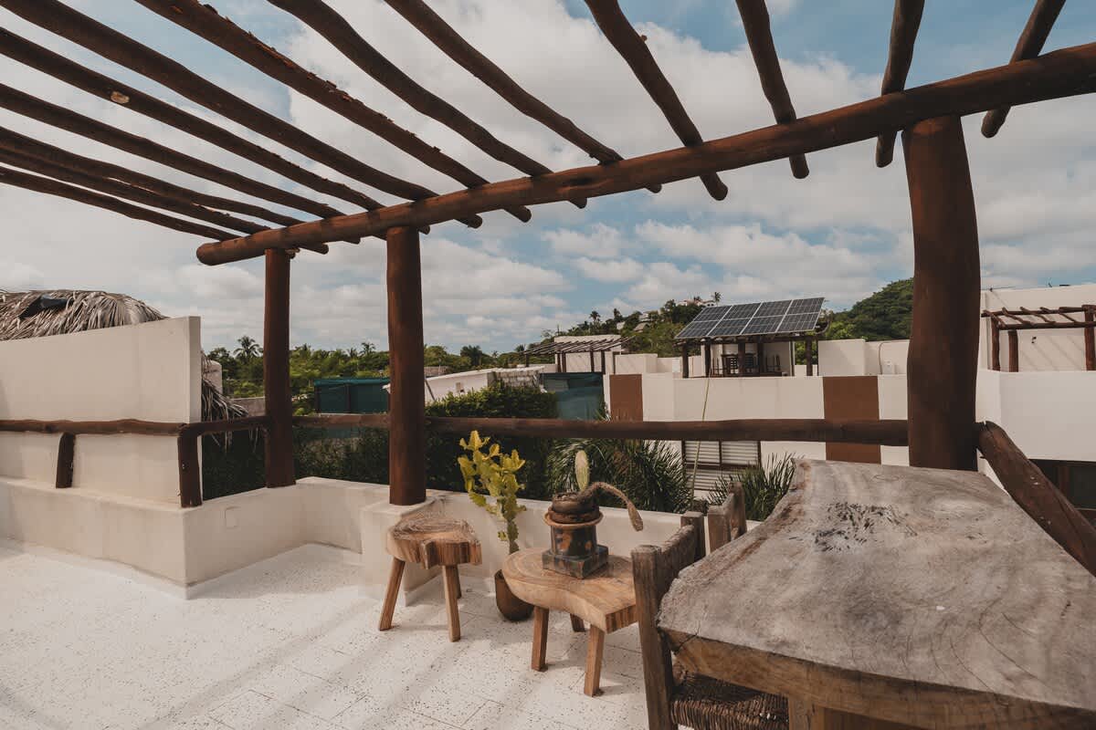 A rustic rooftop deck features a wooden pergola and a high-top bar table with two stools, adorned with a vintage-style water pump decoration, offering views of surrounding buildings and lush tropical foliage.