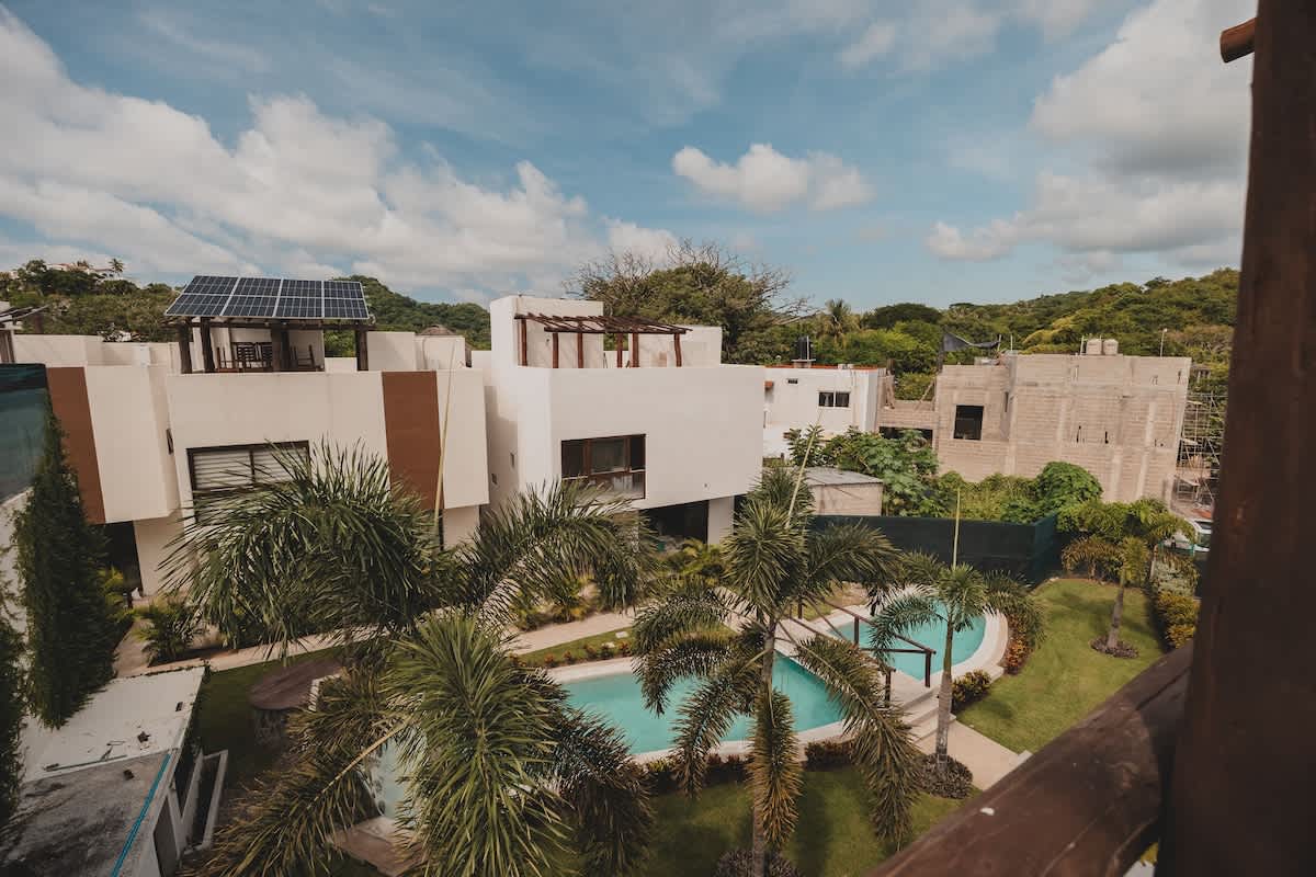 An aerial view showcases a modern white building complex with a winding swimming pool surrounded by palm trees and green lawns, set against a backdrop of lush hills and a partly cloudy sky. Solar panels are visible on one rooftop.