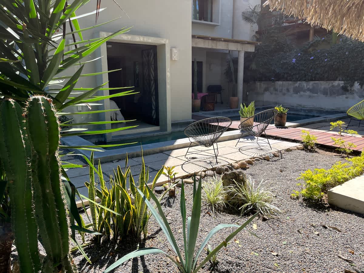 An inviting outdoor space featuring a rectangular swimming pool bordered by concrete pavers and a wooden deck. Lush desert-style landscaping with cacti and agave plants surrounds the area, with black wire chairs providing seating.