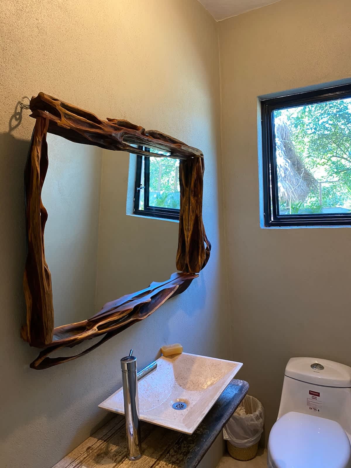 Bathroom featuring a square white sink and a toilet, with a distinctive, organically shaped wooden mirror frame mounted above the sink. Two small windows provide natural light.