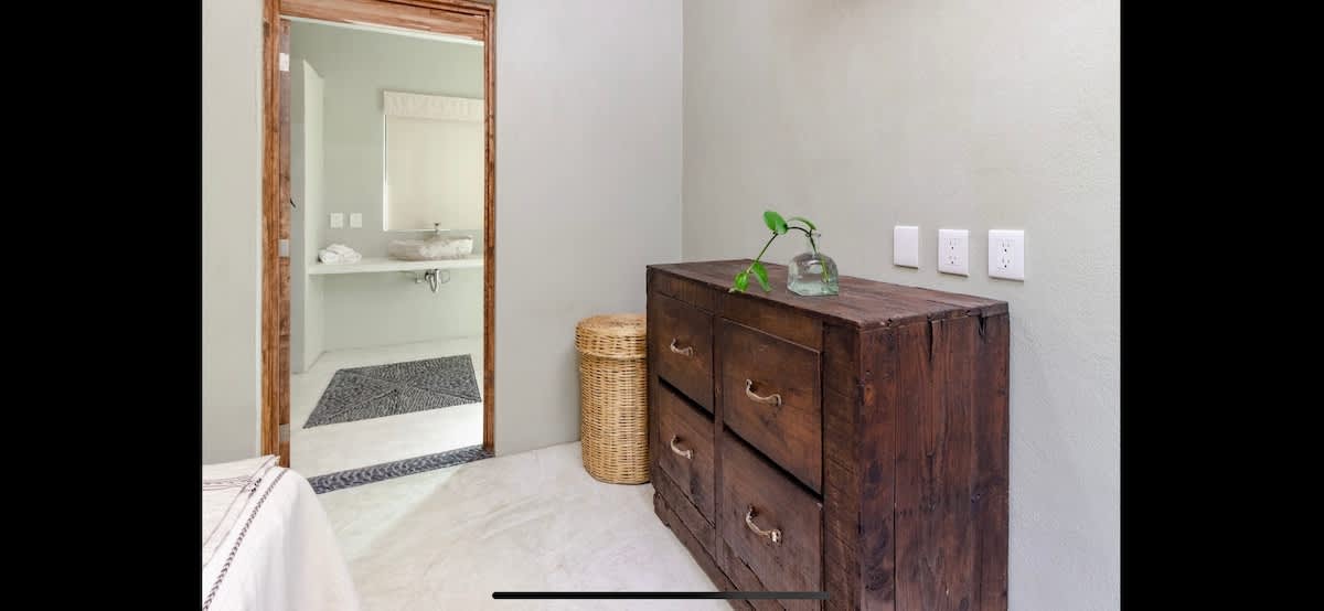 Corner of a light-colored bedroom showing a rustic wooden dresser with a small plant in a vase, a woven basket, and a doorway leading to a bright bathroom with a modern, circular stone sink.