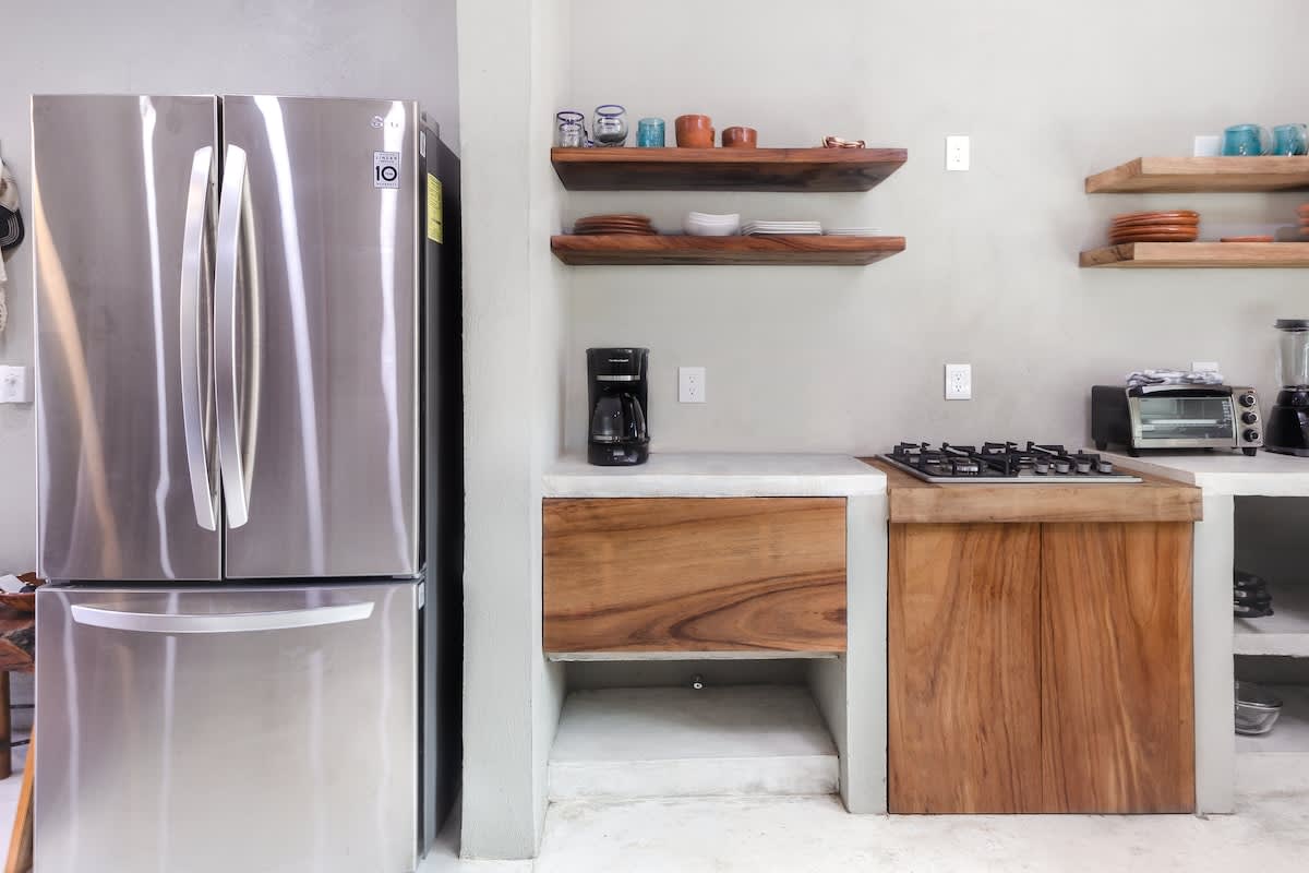 Modern kitchen equipped with a stainless steel French door refrigerator, a gas stovetop integrated into a concrete counter, wooden base cabinets, and open wooden shelves.
