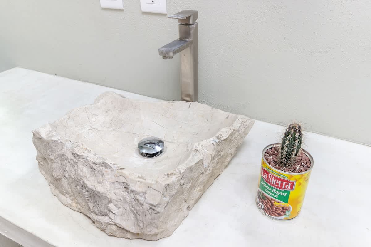 A close-up of a unique, rough-hewn stone vessel sink with a modern brushed metal faucet, set on a light concrete countertop. A small cactus in a repurposed can adds a touch of desert charm.