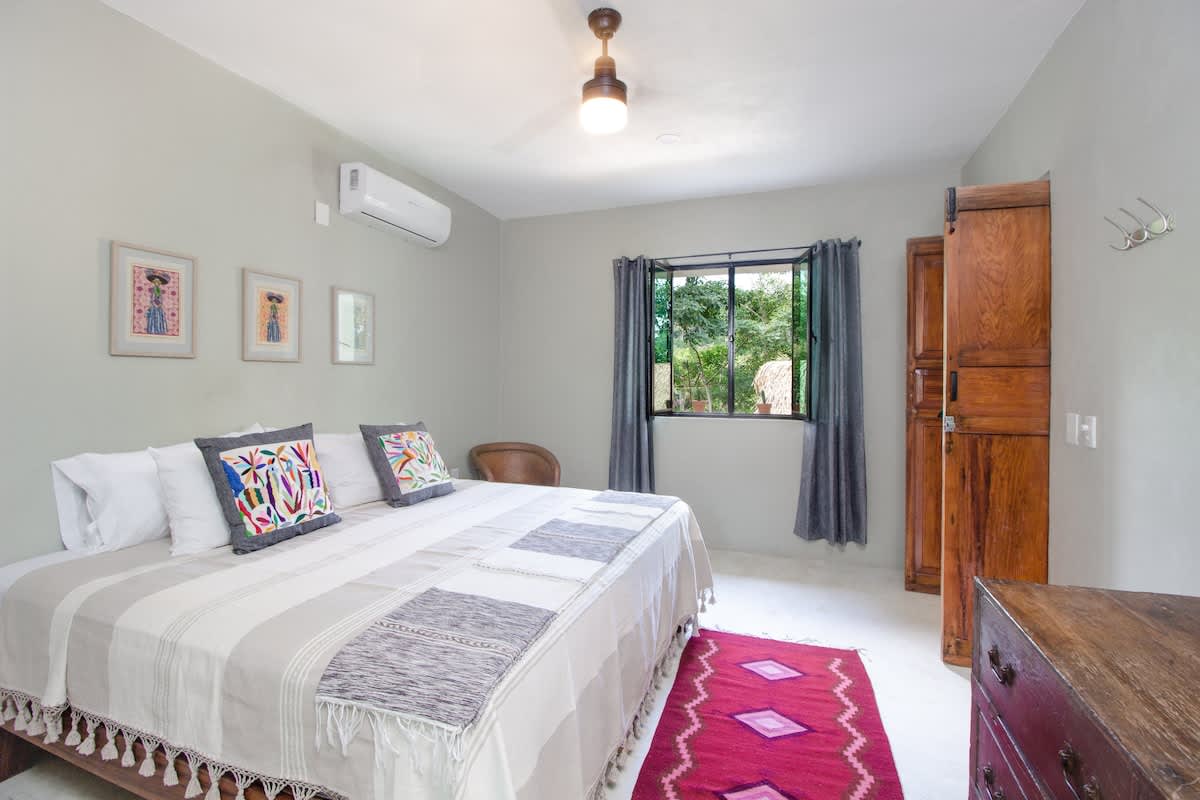 A bright bedroom with light gray walls, a large bed featuring white and striped textiles, and colorful embroidered pillows. A window with dark curtains overlooks lush greenery, and a rustic wooden cabinet stands to the right.