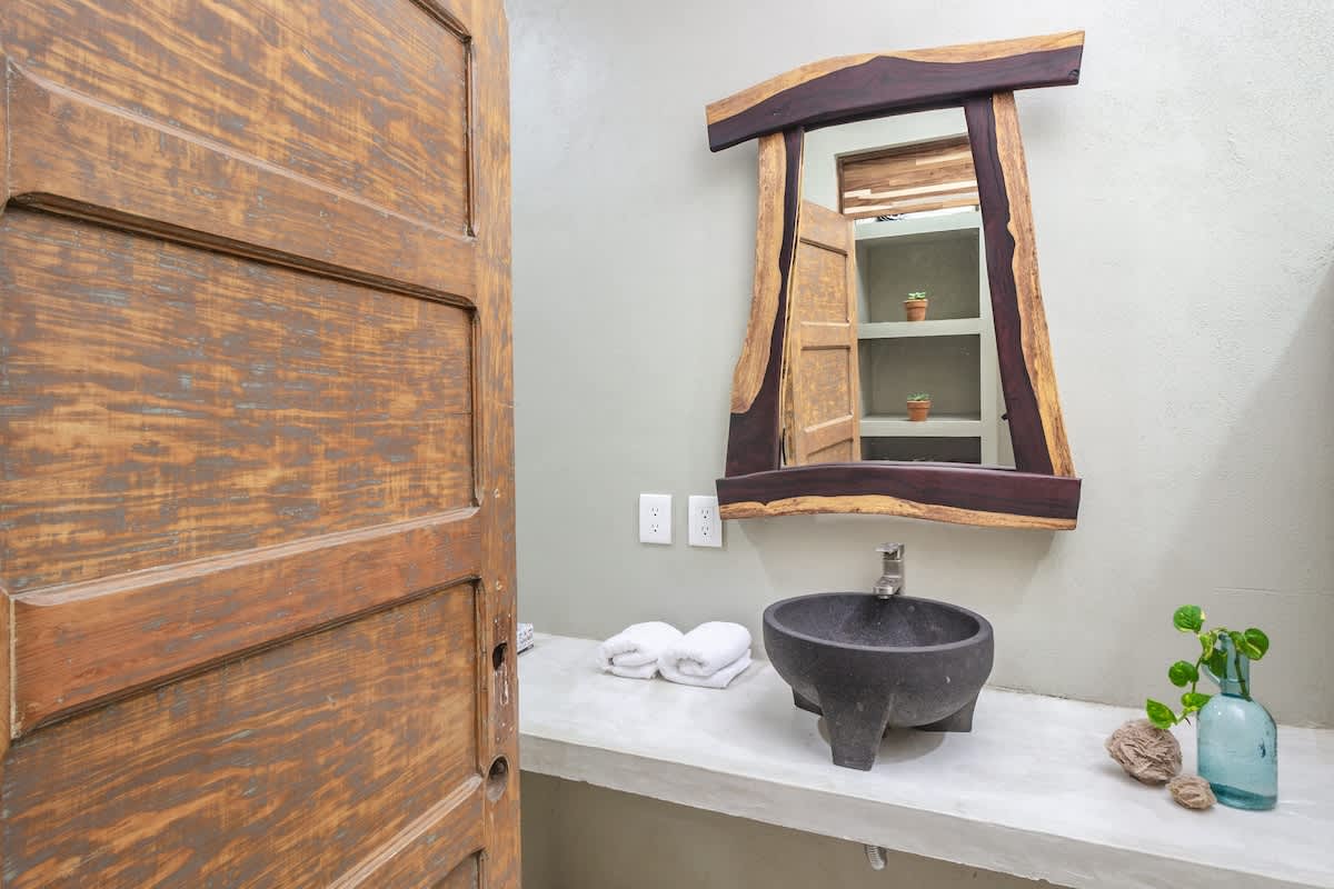 A rustic bathroom featuring a unique live-edge wooden mirror above a concrete vanity with a dark stone vessel sink. A distressed wooden door is visible on the left, adding to the natural aesthetic.