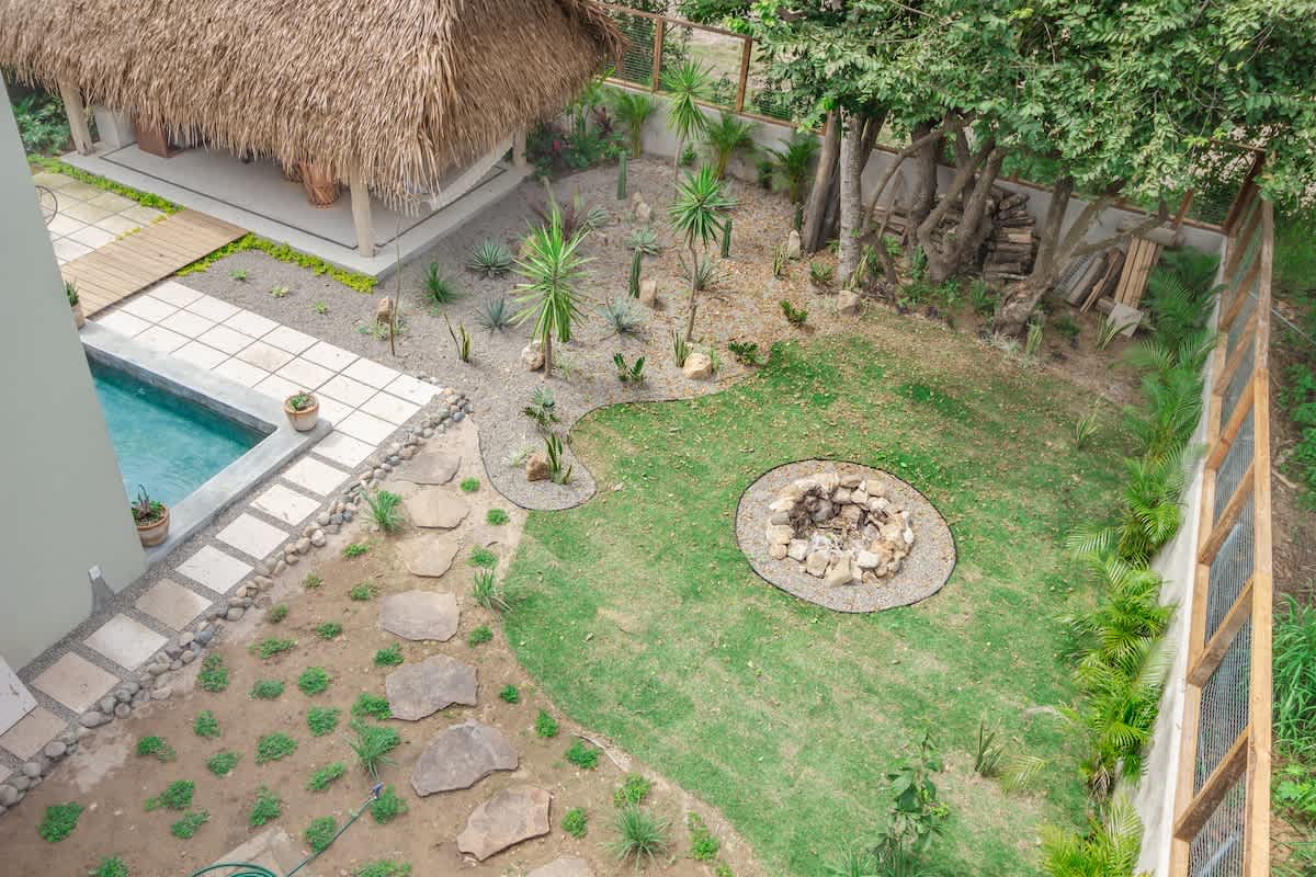 An aerial view of a spacious backyard featuring a rectangular swimming pool, a circular stone fire pit on a grassy lawn, and a large palapa-covered outdoor living area. The garden is landscaped with various plants and trees, enclosed by a fence.