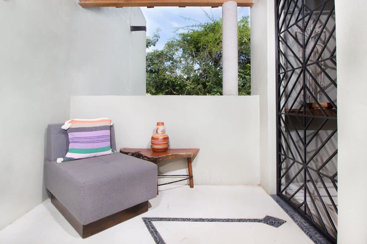 Cozy outdoor seating nook with a grey upholstered armchair, a rustic wooden side table with a decorative vase, and a black ornate metal gate, overlooking greenery.