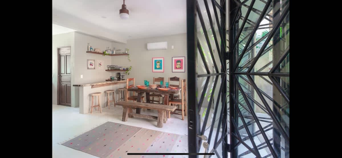 Open-plan dining area with a rustic wooden table and bench seating, adjacent to a bar counter and kitchen, featuring light grey walls, decorative shelves, and large geometric glass doors.