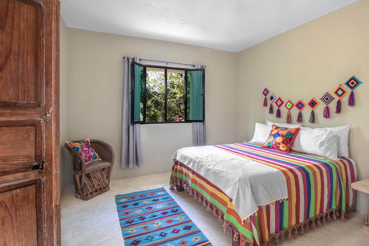 Bright bedroom featuring a bed adorned with a vibrant, striped bedspread and decorative pillows, a small woven chair, a colorful rug, and a window offering a view of lush trees.