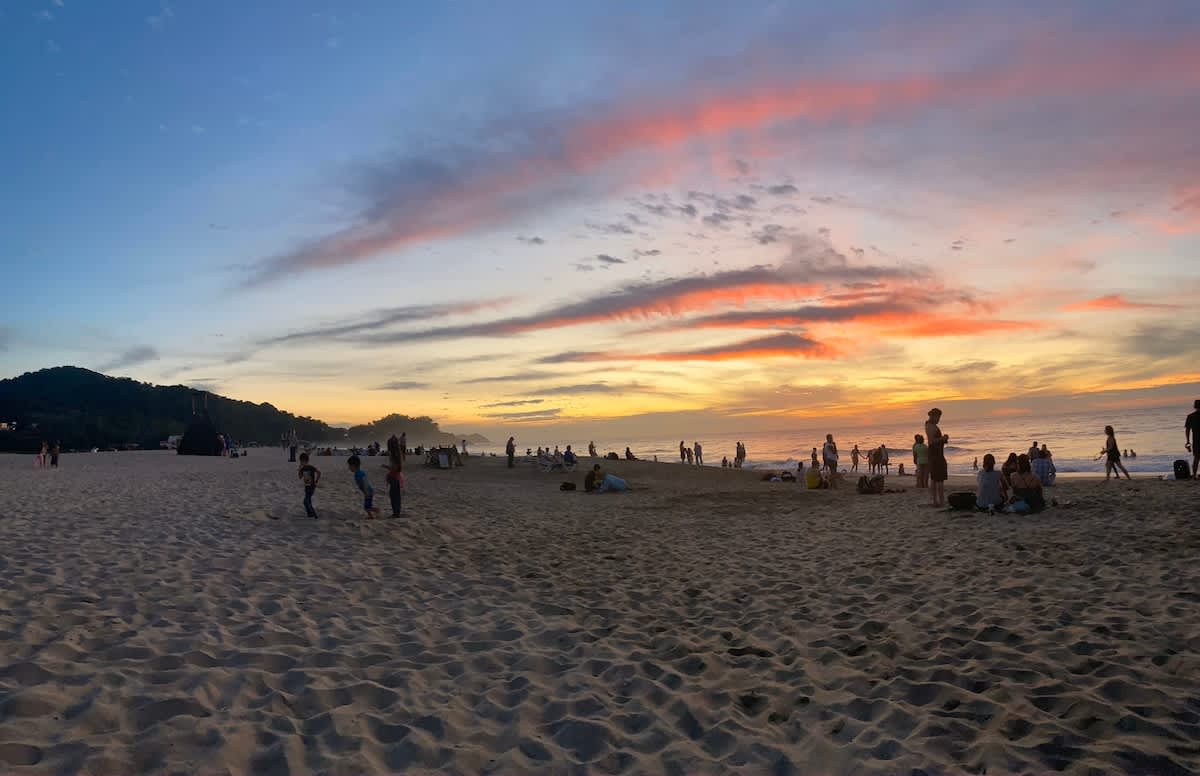 A wide sandy beach at sunset, filled with numerous people enjoying the vibrant orange and pink hues of the sky. Gentle waves lap at the shore, and distant hills frame the scene.