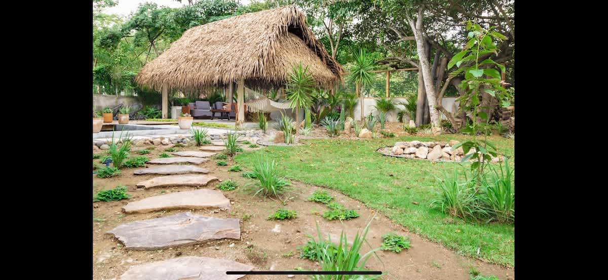 Lush garden pathway made of stepping stones winding through green grass and various plants, leading towards a thatched-roof gazebo with outdoor seating and a hammock in the distance.