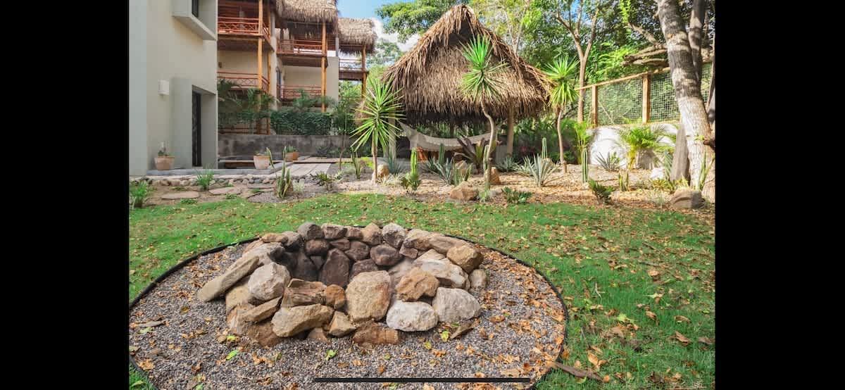 Circular stone fire pit set in a gravel area within a spacious green garden, surrounded by trees and plants, with a thatched-roof structure visible in the background.