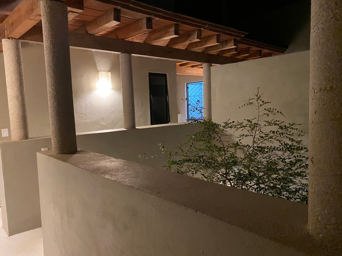 Outdoor covered walkway or balcony at night, featuring sturdy concrete pillars and a wooden beam ceiling, with a wall-mounted light illuminating the path. A small plant adds a touch of greenery in the foreground.