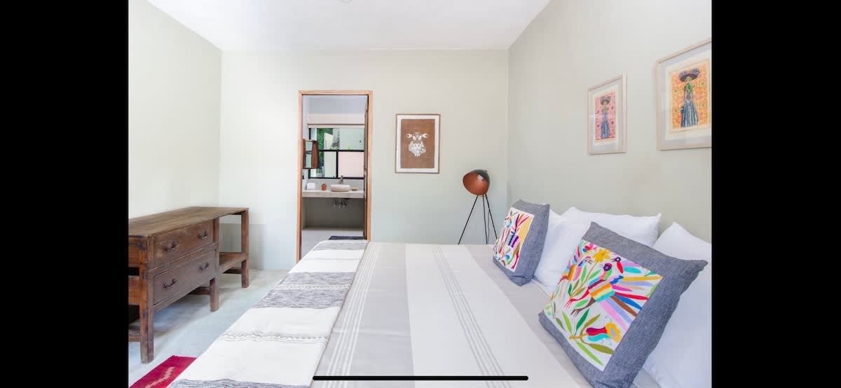 Light-colored bedroom featuring a large bed with white linens and vibrant, embroidered throw pillows, a rustic wooden dresser, and framed artwork on the walls. A doorway leads to an ensuite bathroom with a modern stone sink.