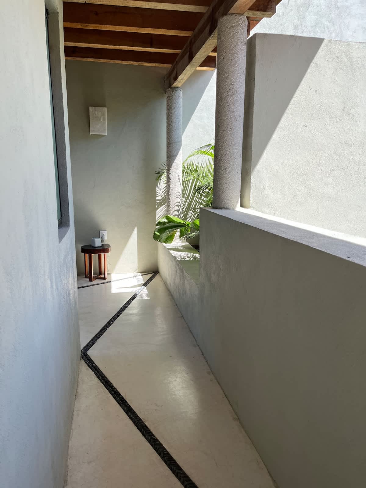 Long, narrow covered outdoor walkway with smooth concrete walls and pillars, a wooden beam ceiling, and a distinctive black and white patterned tile inlay on the floor.
