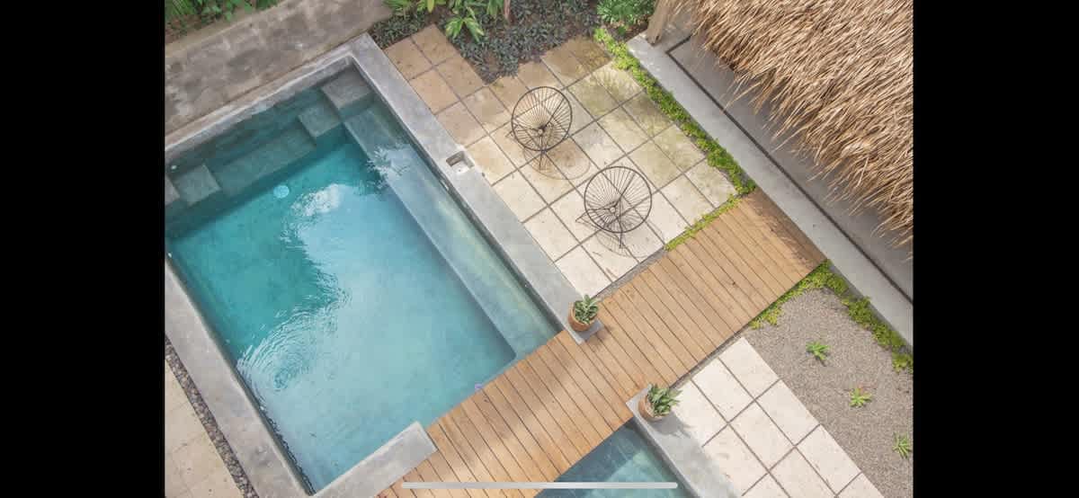 Aerial view of a private pool with clear blue water, surrounded by concrete and stone paving, a wooden walkway, and two wire chairs. A thatched roof structure is visible on the right.