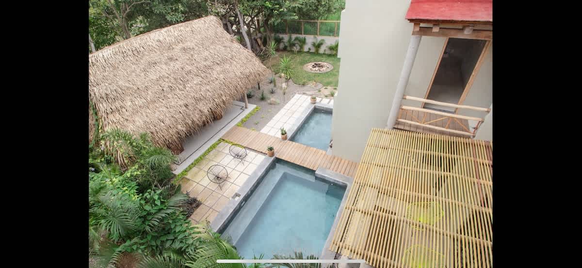 Aerial view of the property's backyard, showcasing two rectangular swimming pools connected by a wooden walkway, surrounded by lush tropical landscaping. Two large thatched-roof palapas provide shade and a resort-like feel.