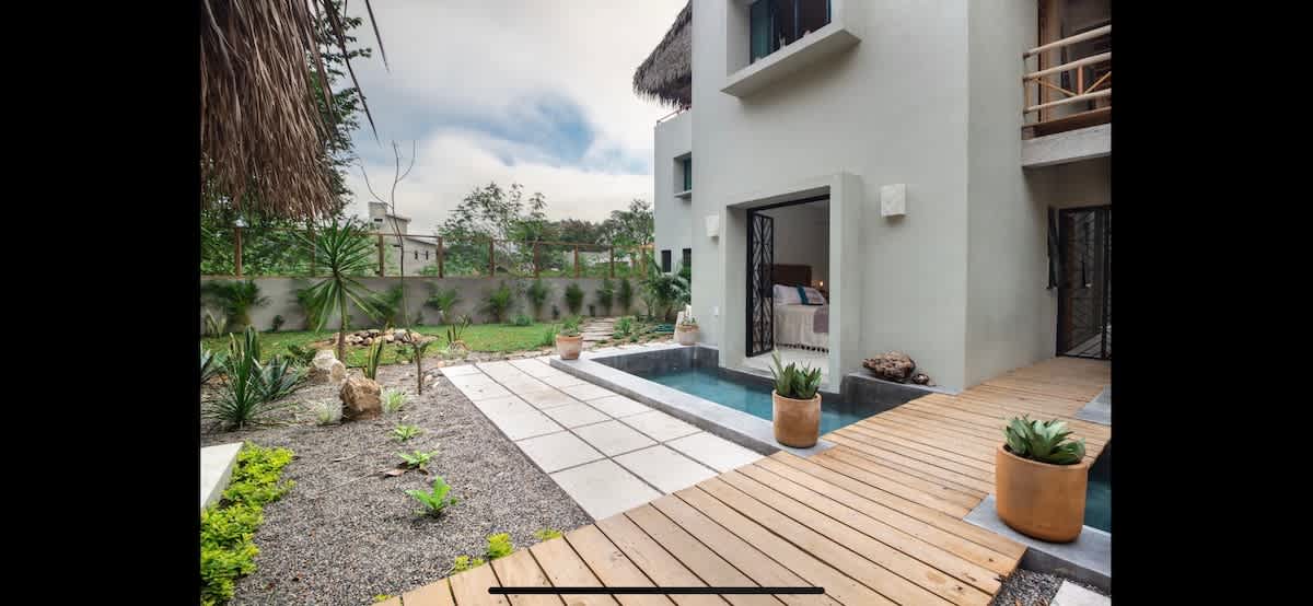 Modern building exterior with light grey walls and a thatched roof, overlooking a small pool with a wooden deck and a landscaped garden featuring various desert plants and gravel pathways.