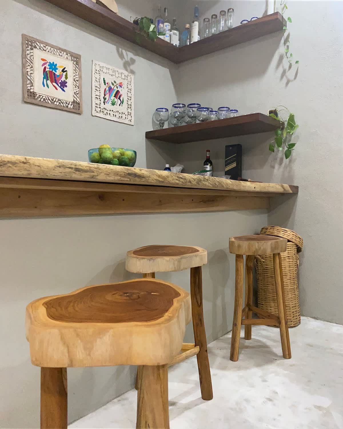 Rustic bar area featuring a live-edge wooden counter and three matching wooden stools. Shelves above display bottles and glassware, with framed artwork on the light gray wall.