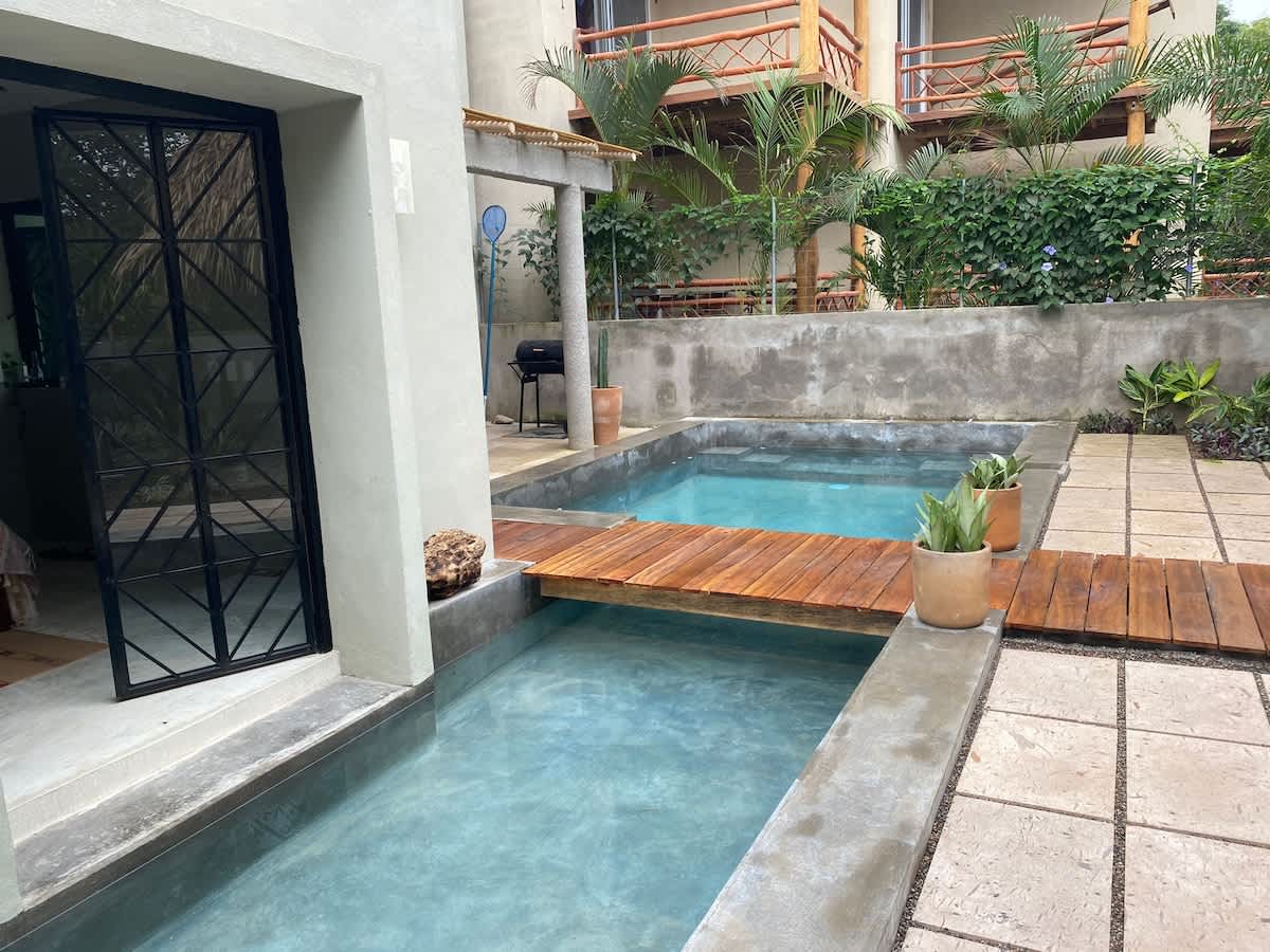 Outdoor patio area featuring two small, rectangular plunge pools connected by a wooden bridge, with a concrete surround and a BBQ grill visible in the background.
