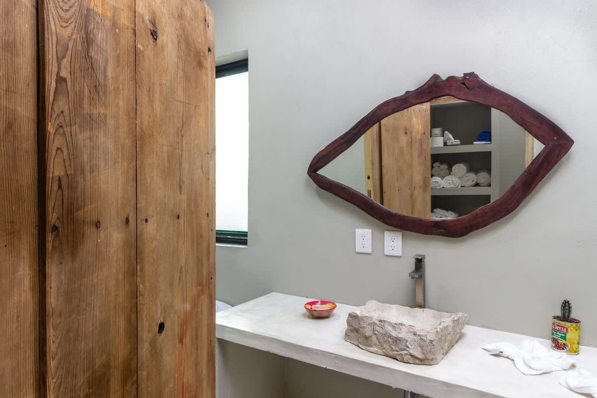 A rustic bathroom with a unique, eye-shaped wooden mirror above a concrete vanity. A large, rough-hewn stone vessel sink is featured, and a distressed wooden wall or door is visible to the left.