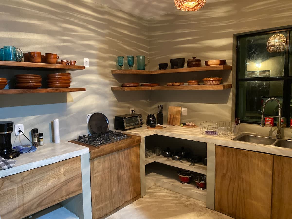 Rustic kitchen with light concrete countertops and wooden base cabinets, featuring open wooden shelving displaying various pottery and glassware. A gas stovetop and a double sink are visible under a large window.
