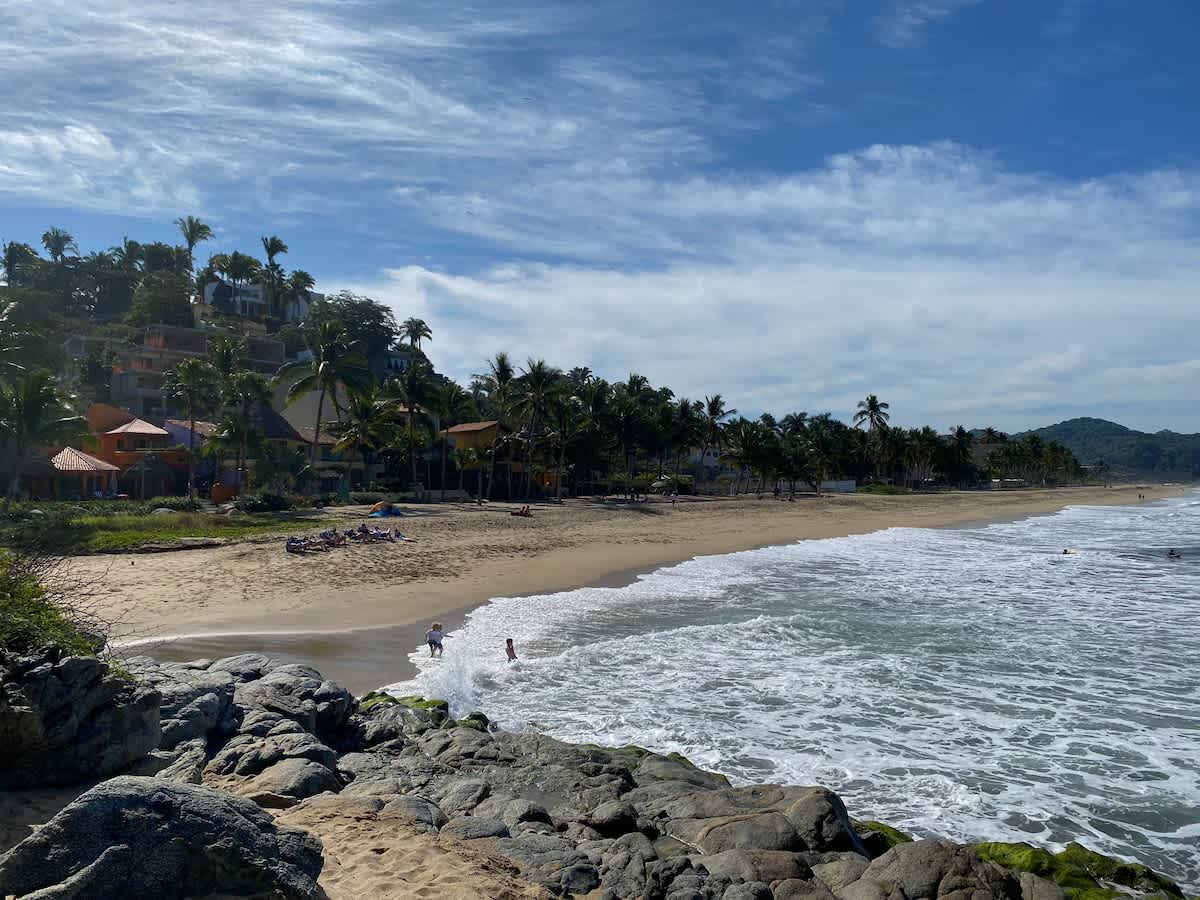 Expansive view of a sandy beach with crashing waves and large rocks in the foreground. Palm trees and houses dot the hillside in the background under a blue sky with white clouds.