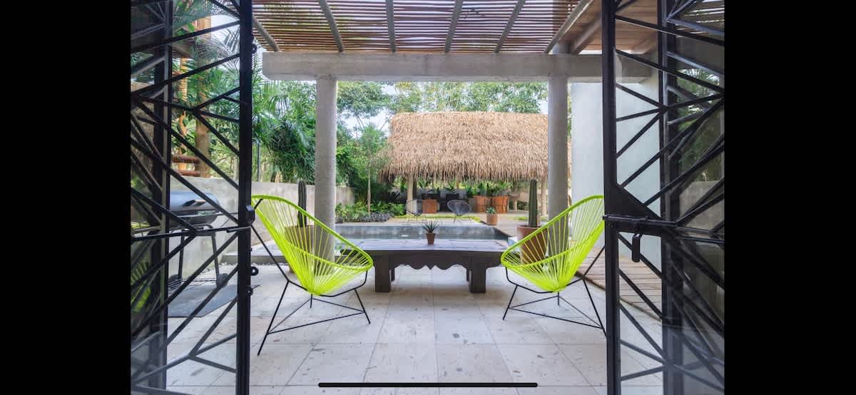 Outdoor patio area viewed through decorative black metal gates, featuring two bright neon green Acapulco chairs and a rustic wooden coffee table. A large thatched-roof palapa and lush tropical greenery are visible in the background.
