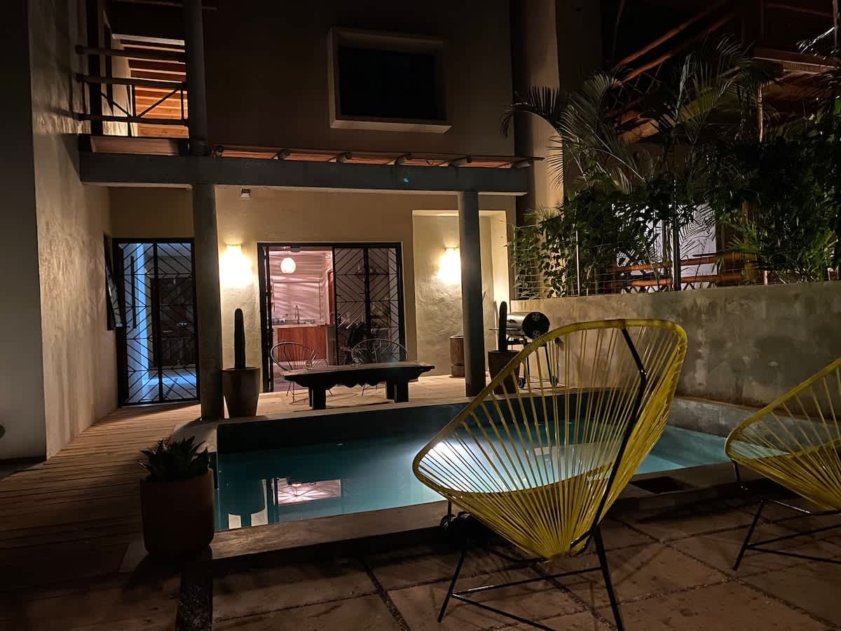 Nighttime view of an outdoor area with a small, illuminated pool and two bright yellow Acapulco chairs. The surrounding buildings are softly lit, creating a warm and inviting ambiance.
