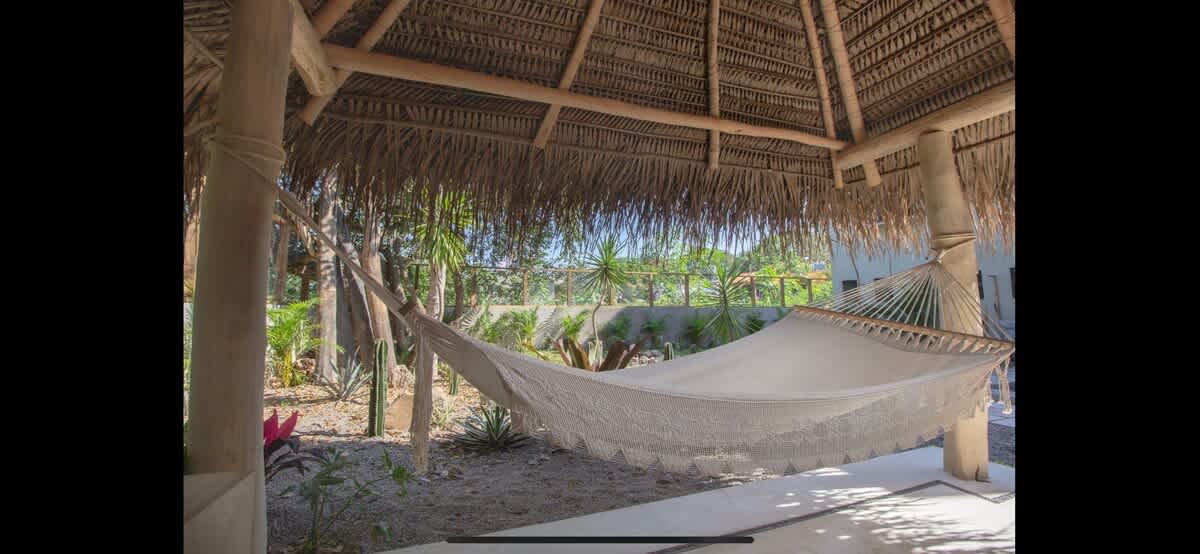 Inviting white woven hammock suspended under a rustic thatched palapa roof, surrounded by lush tropical foliage, offering a serene spot for relaxation.