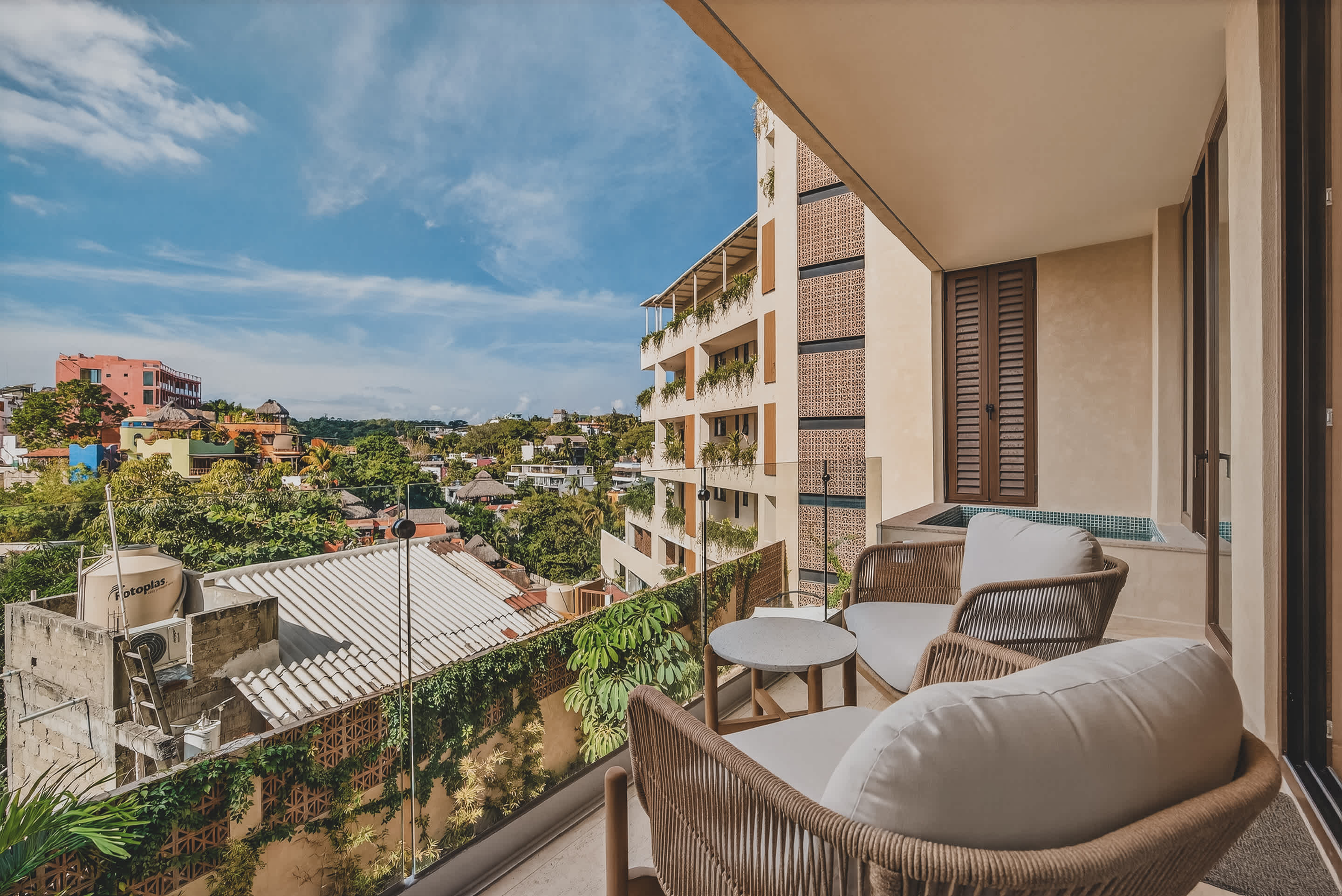 A spacious balcony features two woven armchairs with white cushions and a small round table, overlooking a vibrant town with lush trees and buildings. A private plunge pool is visible to the left, and a modern building facade is across the way.
