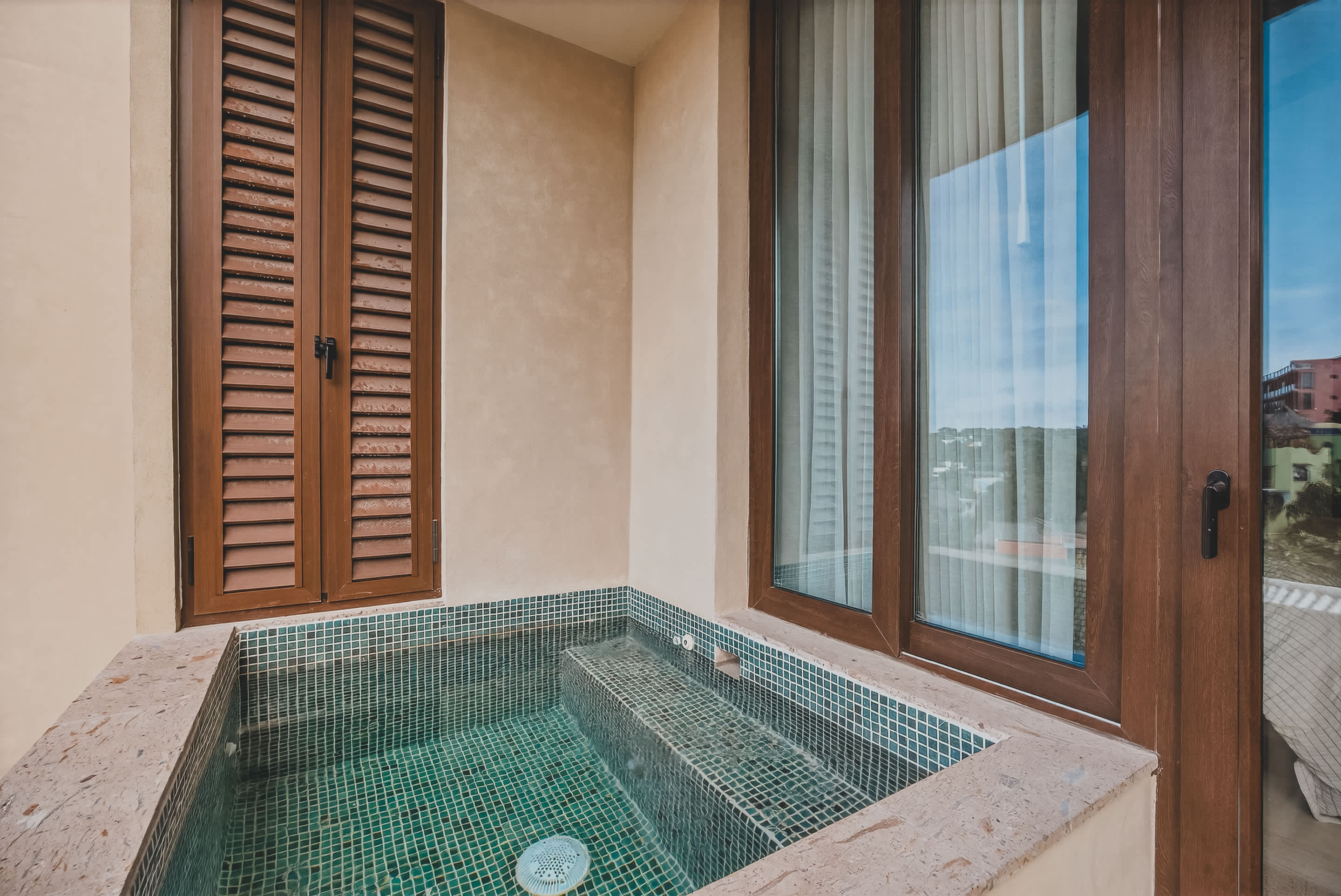 A private balcony features a small tiled plunge pool with steps, adjacent to large glass sliding doors that open to an interior space. Brown louvered shutters are on the left, and the pool is surrounded by light-colored stone coping.