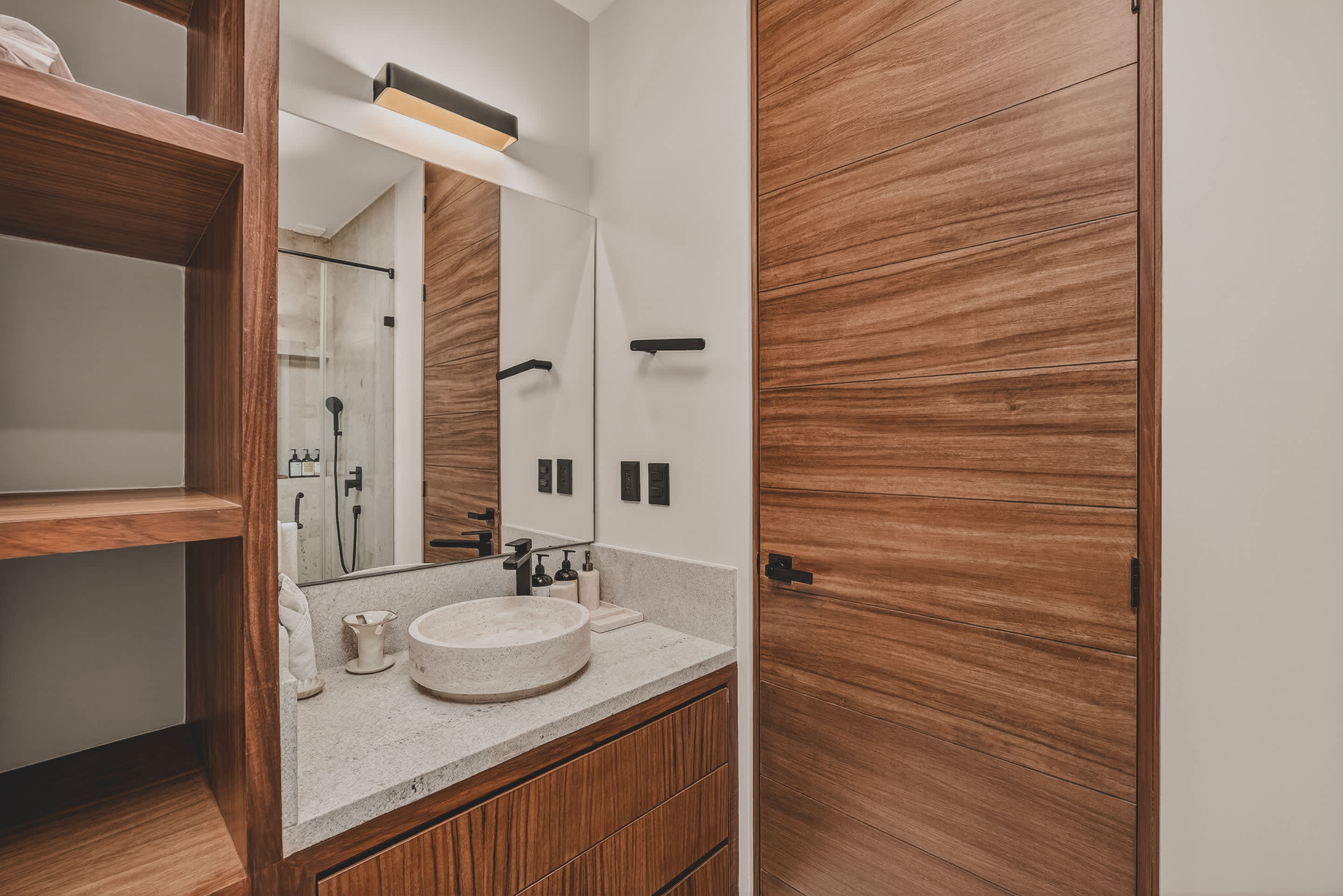 A contemporary bathroom features a light stone vanity with a round vessel sink and black faucet, integrated with open wooden shelving. A large mirror reflects a glass-enclosed shower and a wooden door.