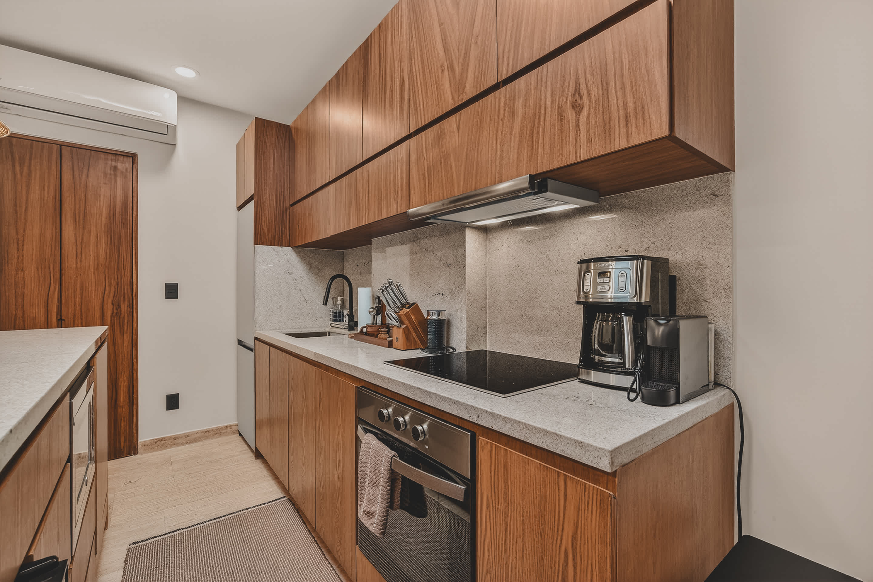 A well-equipped kitchen counter features a black induction cooktop, a coffee maker, and a toaster, all set against a light speckled backsplash and dark wood cabinetry. An oven is integrated below the cooktop, with a towel hanging from its handle.