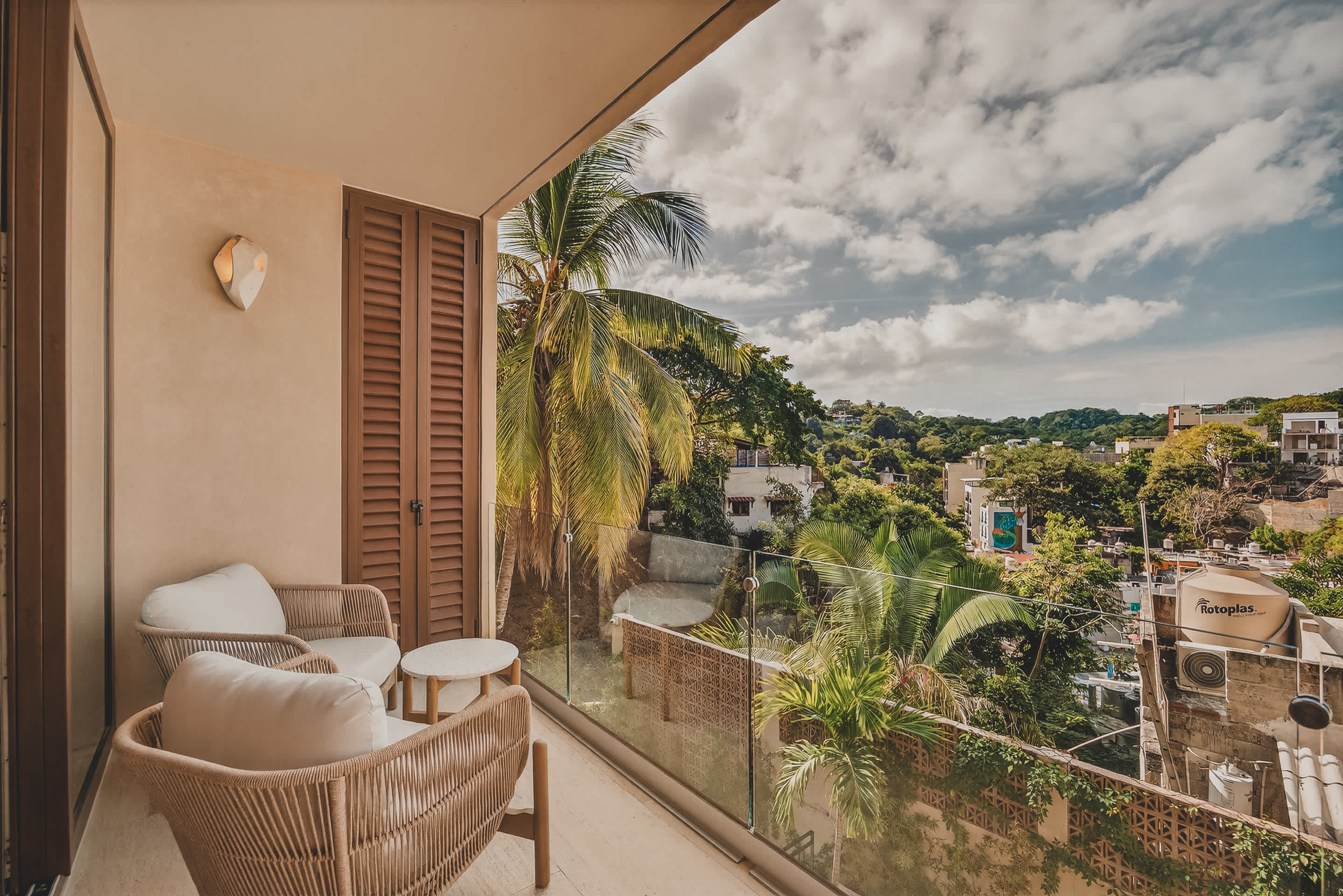 Cozy balcony furnished with two rattan armchairs and a small round side table, offering a scenic view of palm trees and distant buildings under a partly cloudy sky.