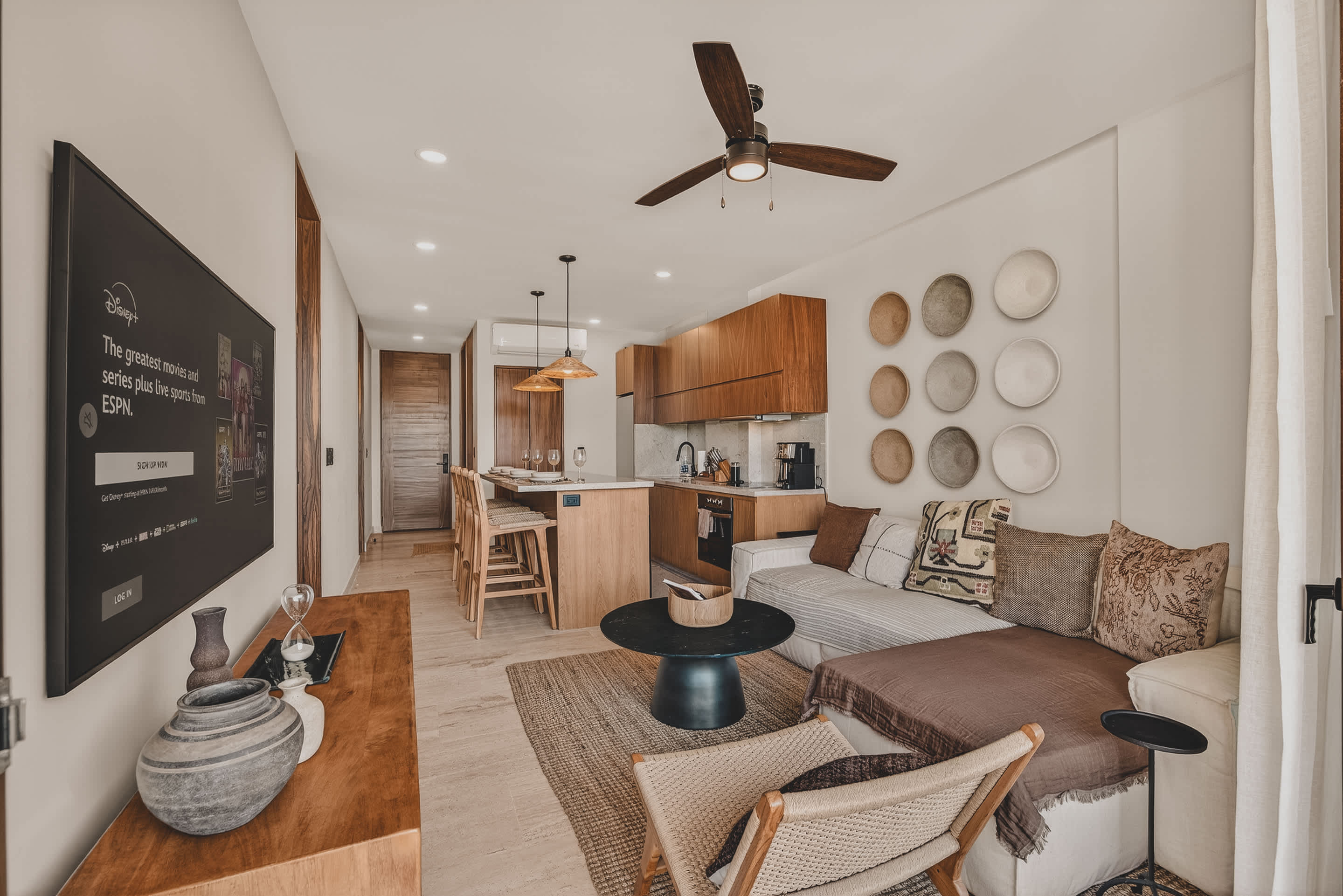 An inviting open-concept living space combines a comfortable L-shaped sofa with a modern kitchen and dining island, all featuring warm wood tones and neutral decor. A large wall-mounted TV and decorative wall plates add character, with a glimpse of a bedroom through a doorway.