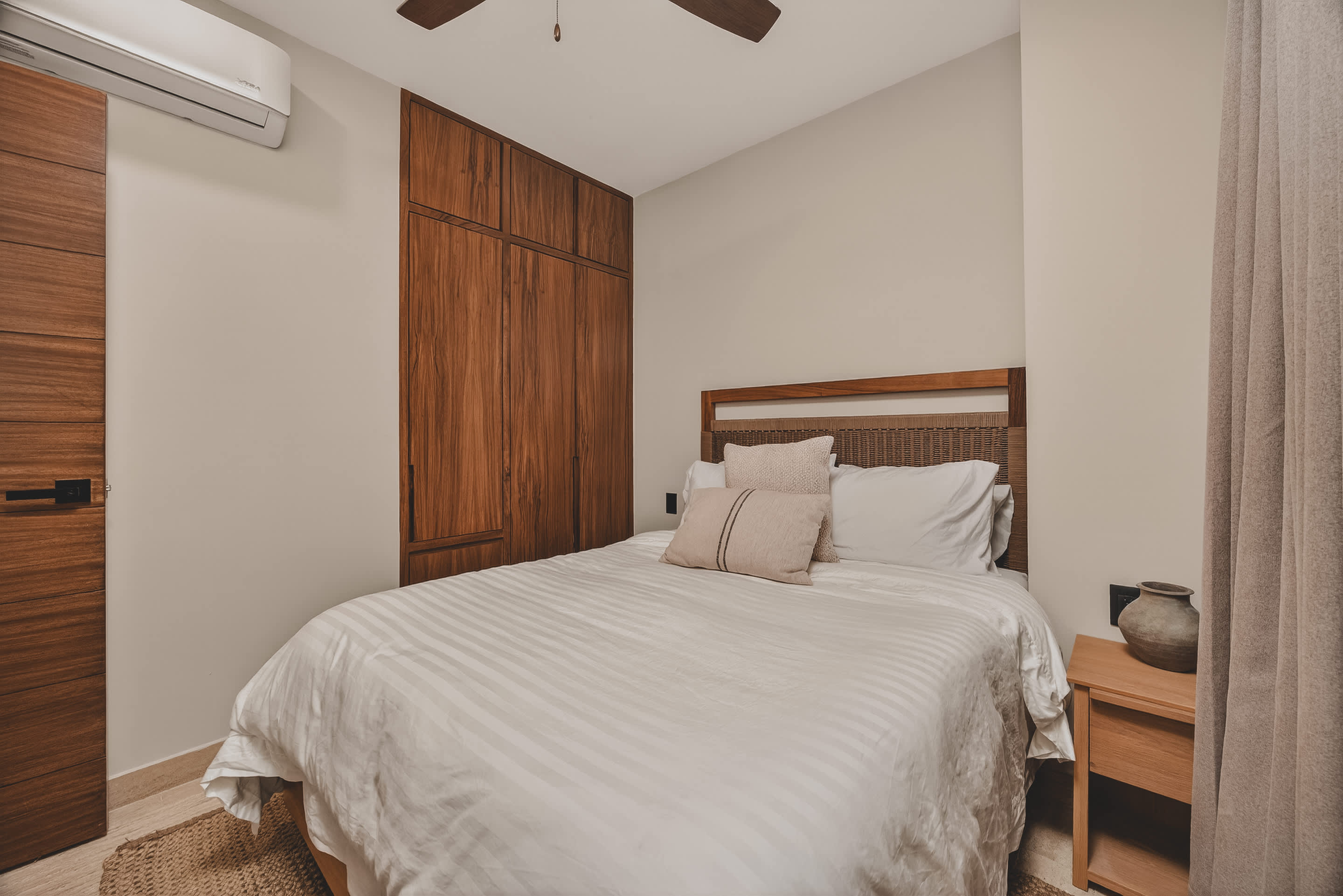 A cozy bedroom features a queen-sized bed with a woven headboard and light-colored bedding, flanked by a wooden nightstand. A large built-in wooden wardrobe spans one wall, and an air conditioning unit is mounted above the door.