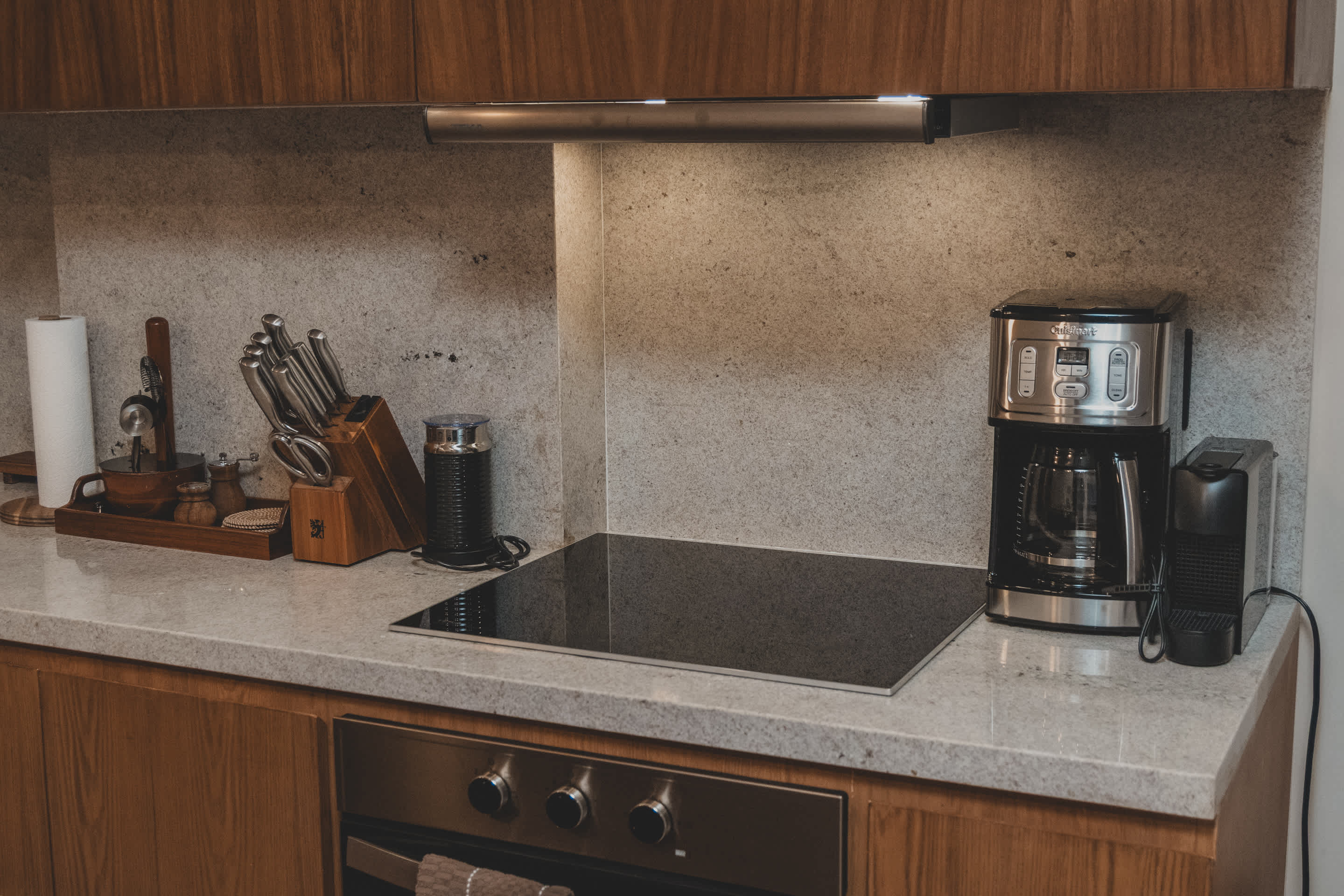 A well-equipped kitchen counter features a black induction cooktop, a coffee maker, a knife block, and a Nespresso machine, all set against a light speckled granite backsplash and wooden cabinetry.