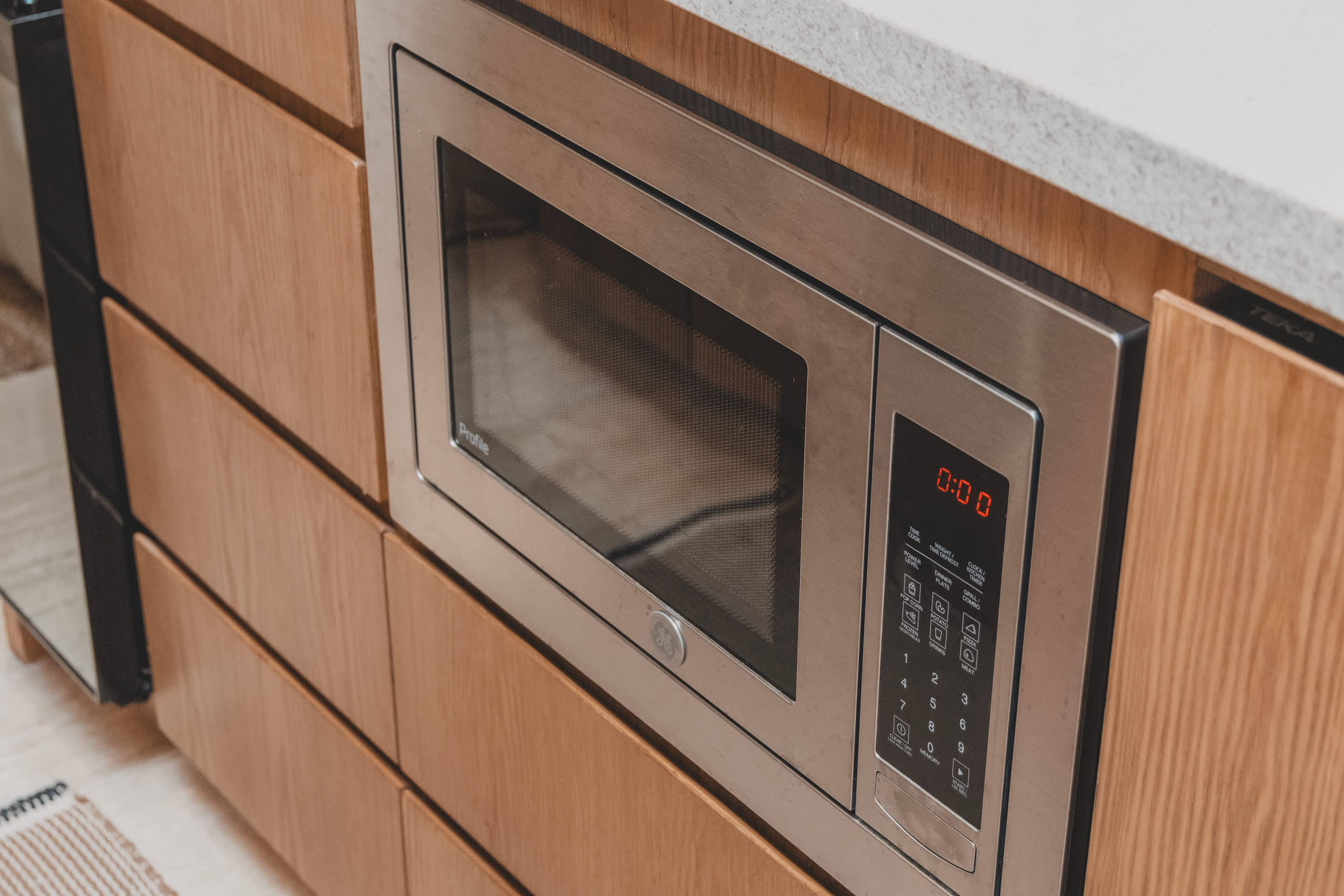 A stainless steel built-in microwave with a digital display showing "0:00" is seamlessly integrated into light wooden kitchen cabinetry.