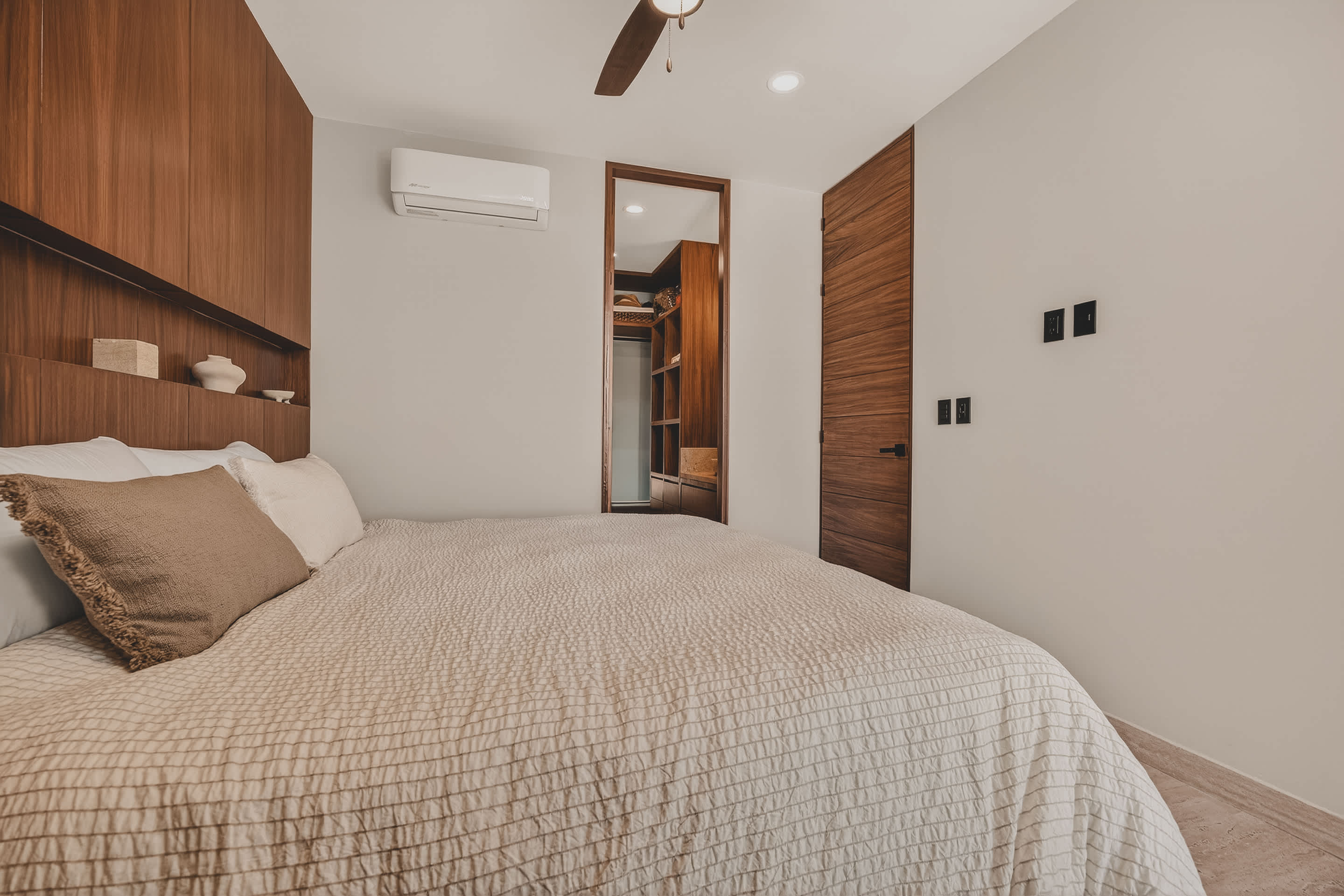 A modern bedroom features a king-sized bed with light bedding, positioned against a dark wooden accent wall with integrated shelving. An air conditioning unit is mounted above a doorway leading to a walk-in closet or bathroom.