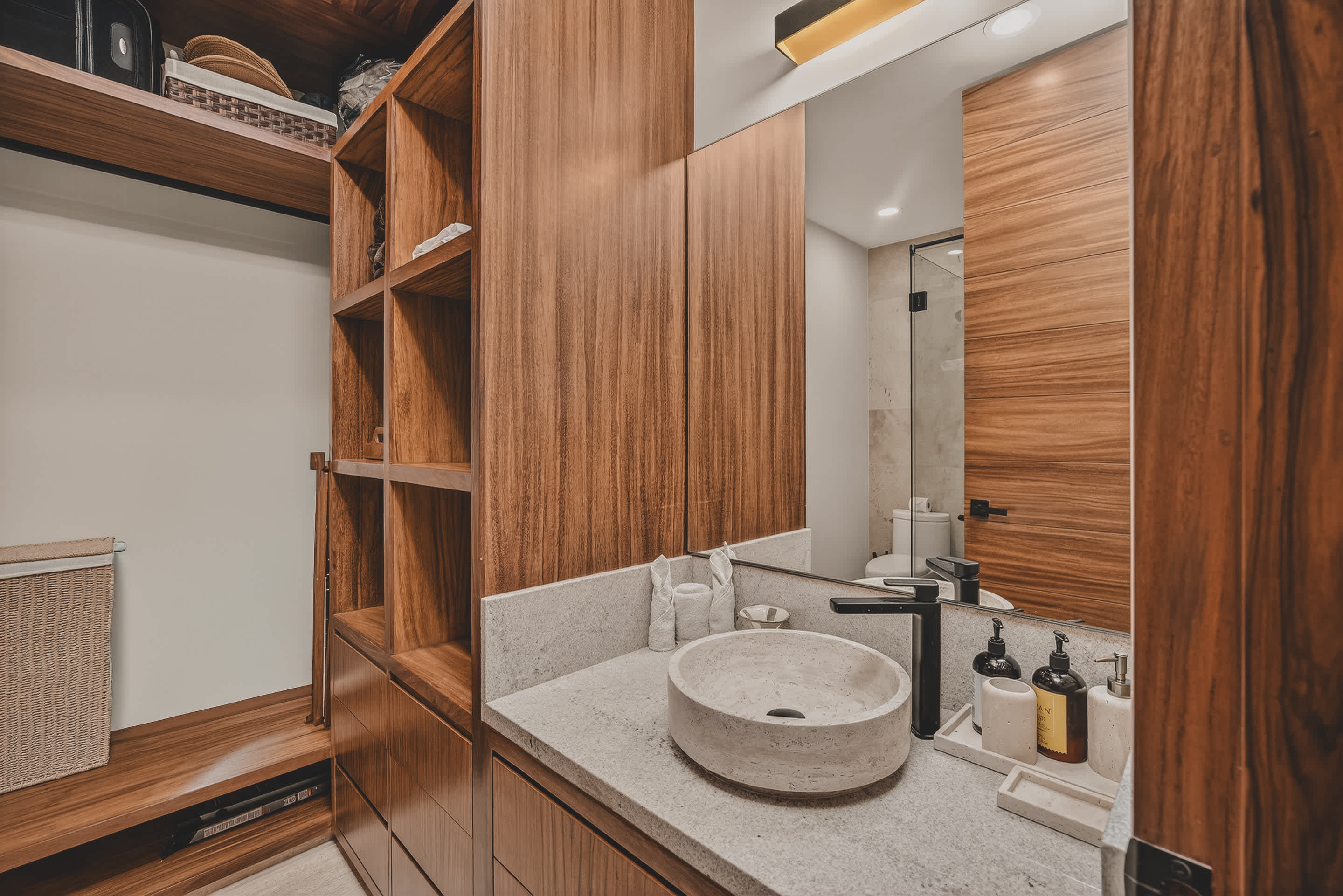 A modern bathroom vanity area features a light stone countertop with a round vessel sink and black fixtures, alongside open wooden shelving. A large mirror reflects a shower area and a wooden door.