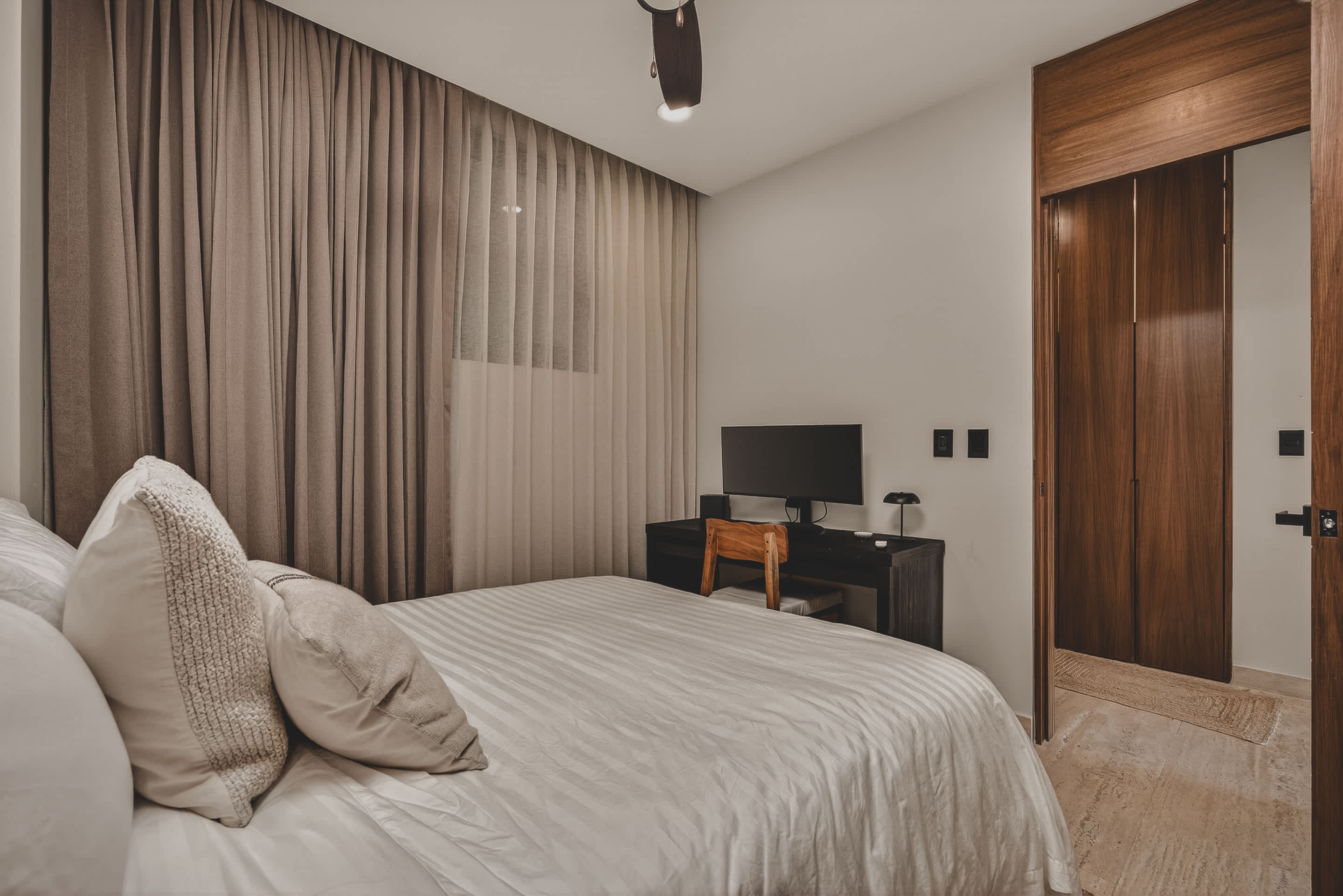 Another view of the bedroom showing the queen-sized bed with a rattan headboard, a dark wooden desk with a monitor, and floor-to-ceiling curtains, with a doorway visible to the right.