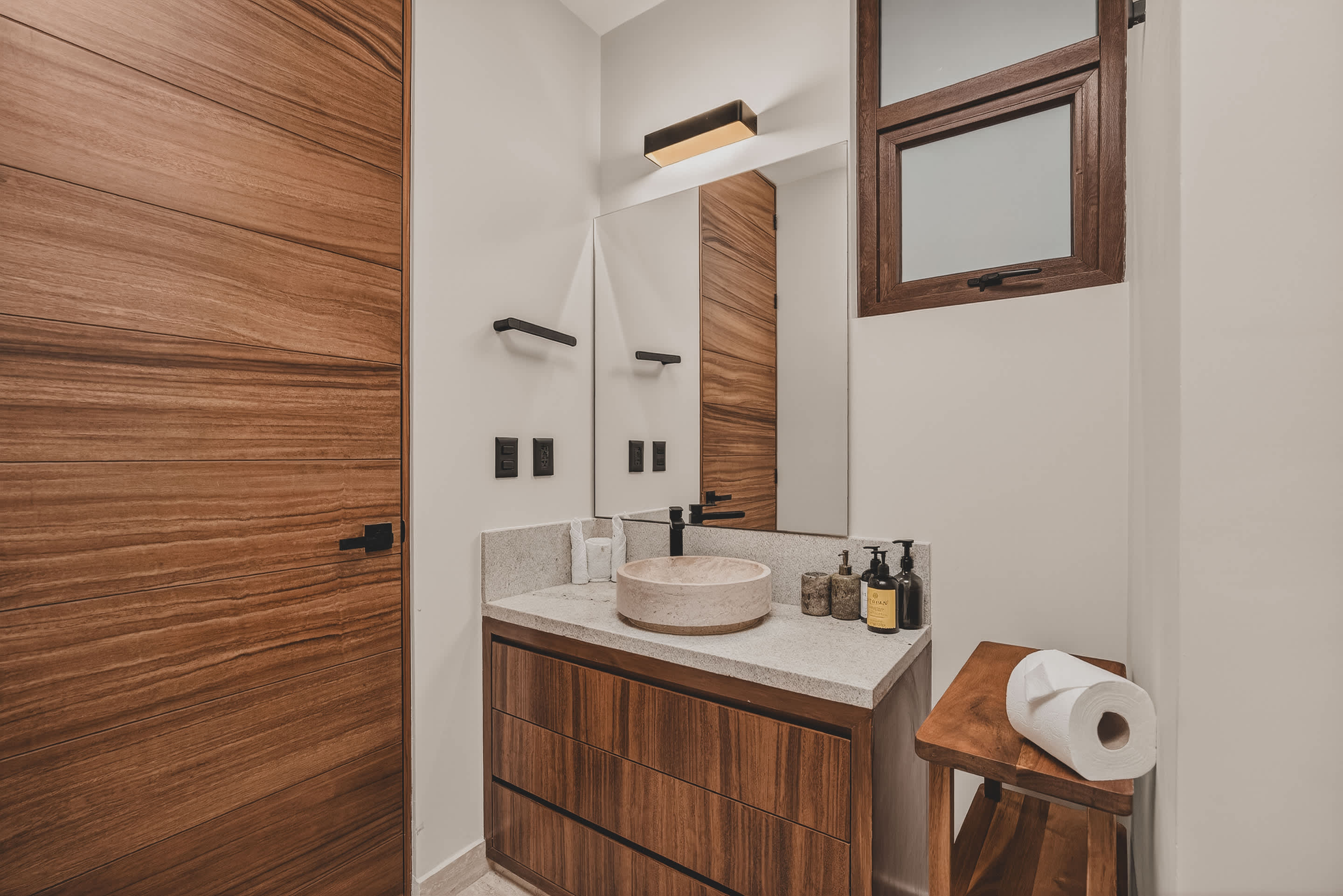 Another perspective of the modern bathroom, highlighting the wooden vanity with a vessel sink, black fixtures, and a large mirror, with a window above a wooden shelf.