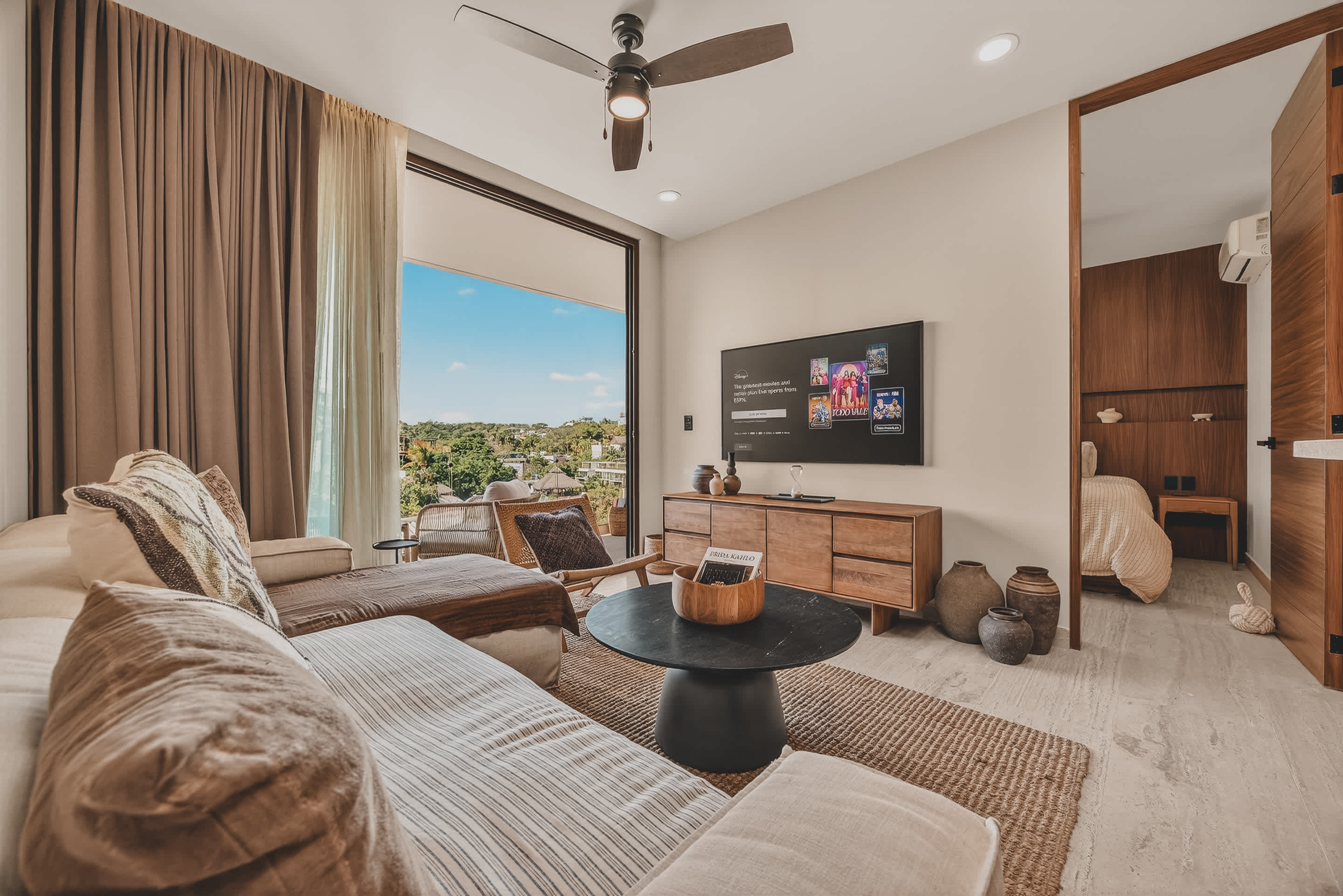 A bright living room features a large L-shaped sofa with throw pillows, a round black coffee table, and a wall-mounted TV displaying streaming content. Sliding glass doors open to a balcony with a scenic view, and a bedroom is visible through an open wooden doorway.
