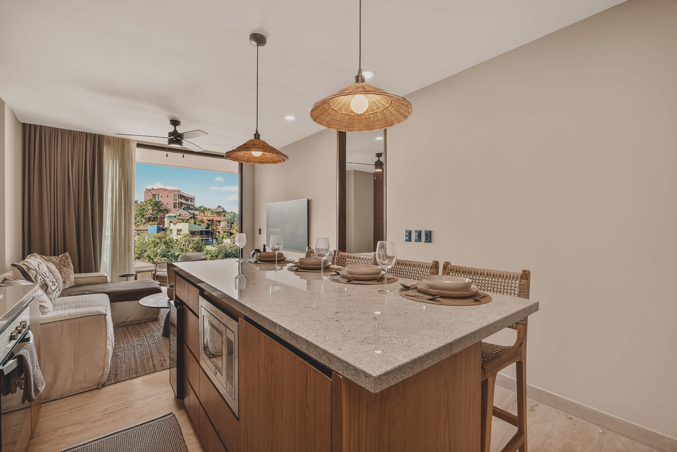 A spacious kitchen island with a light speckled countertop and four woven bar stools serves as a dining area, with a microwave integrated below. The open layout connects to a living room with a sofa and a balcony offering an outdoor view.