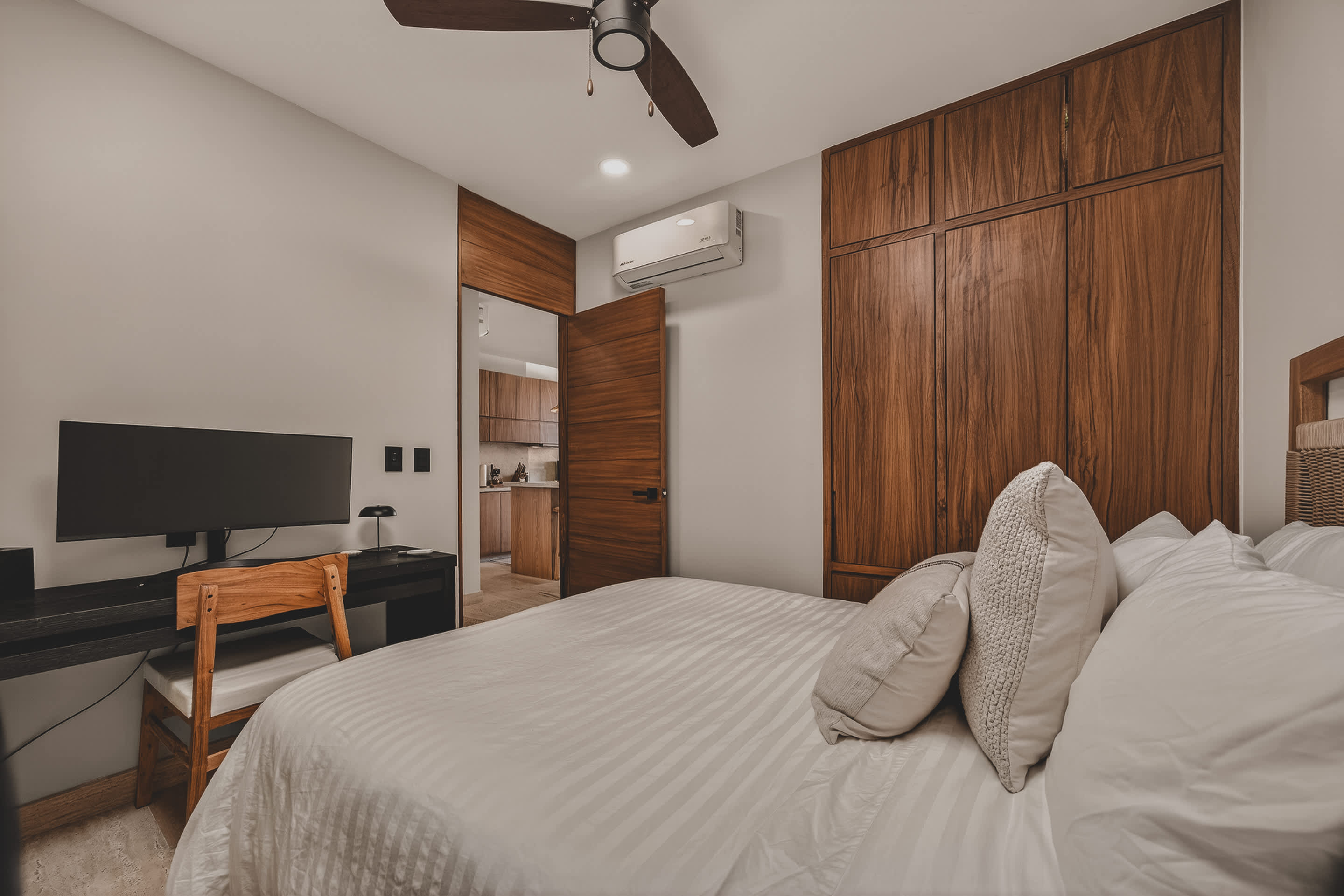 Close-up of the bedroom's desk area with a monitor and a wooden chair, showing the built-in wooden wardrobe and a doorway leading to another room, possibly the kitchen or hallway.