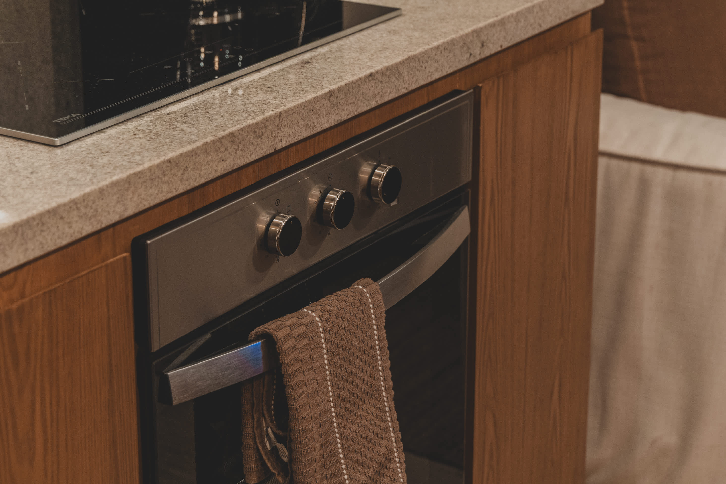 A stainless steel built-in oven with four control knobs is integrated into wooden cabinetry, with a brown striped towel hanging from its handle.