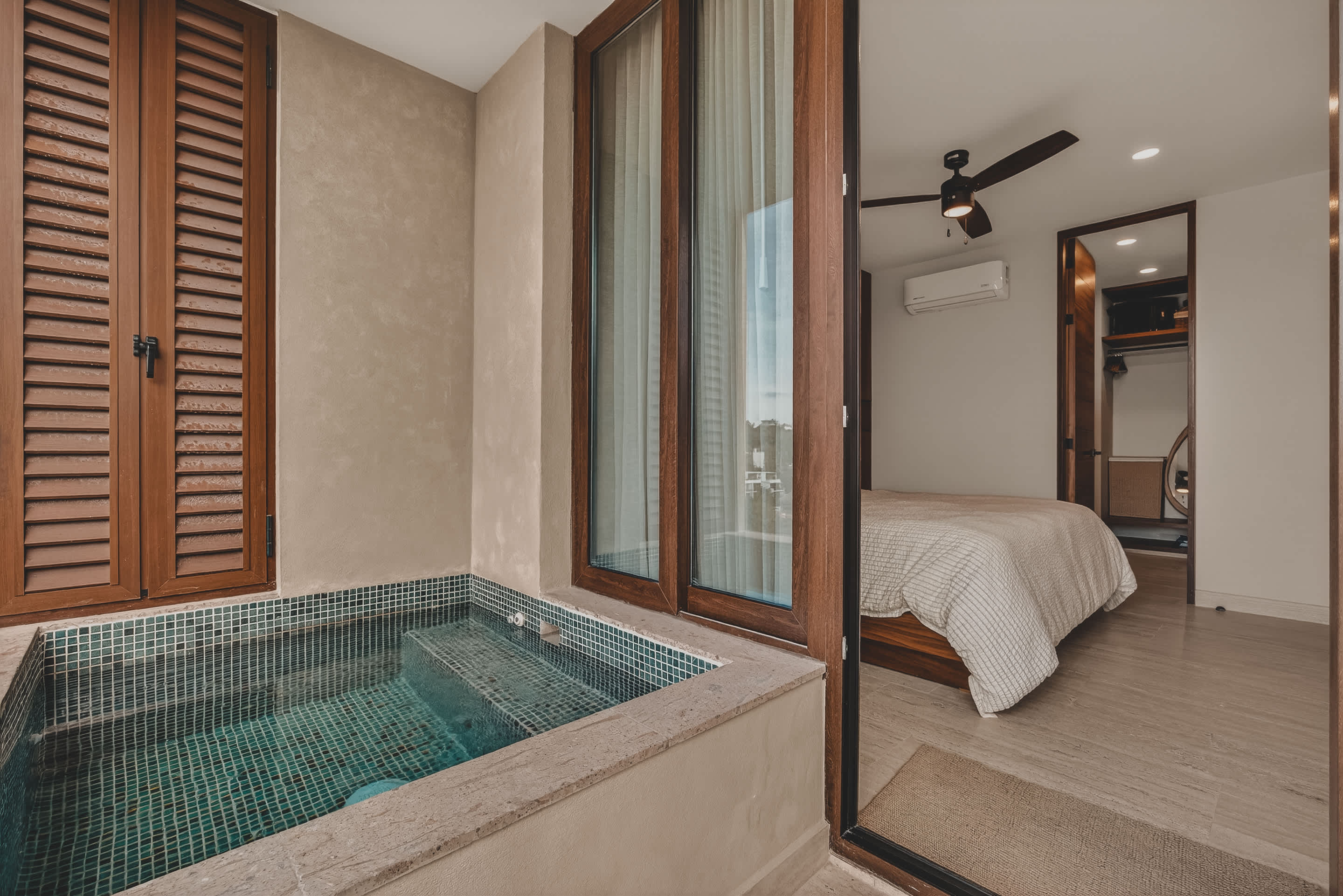 A private balcony with a tiled plunge pool is seen from inside, with large glass doors opening to a bedroom featuring a bed with light bedding and a ceiling fan. The bedroom has a modern wooden accent wall.