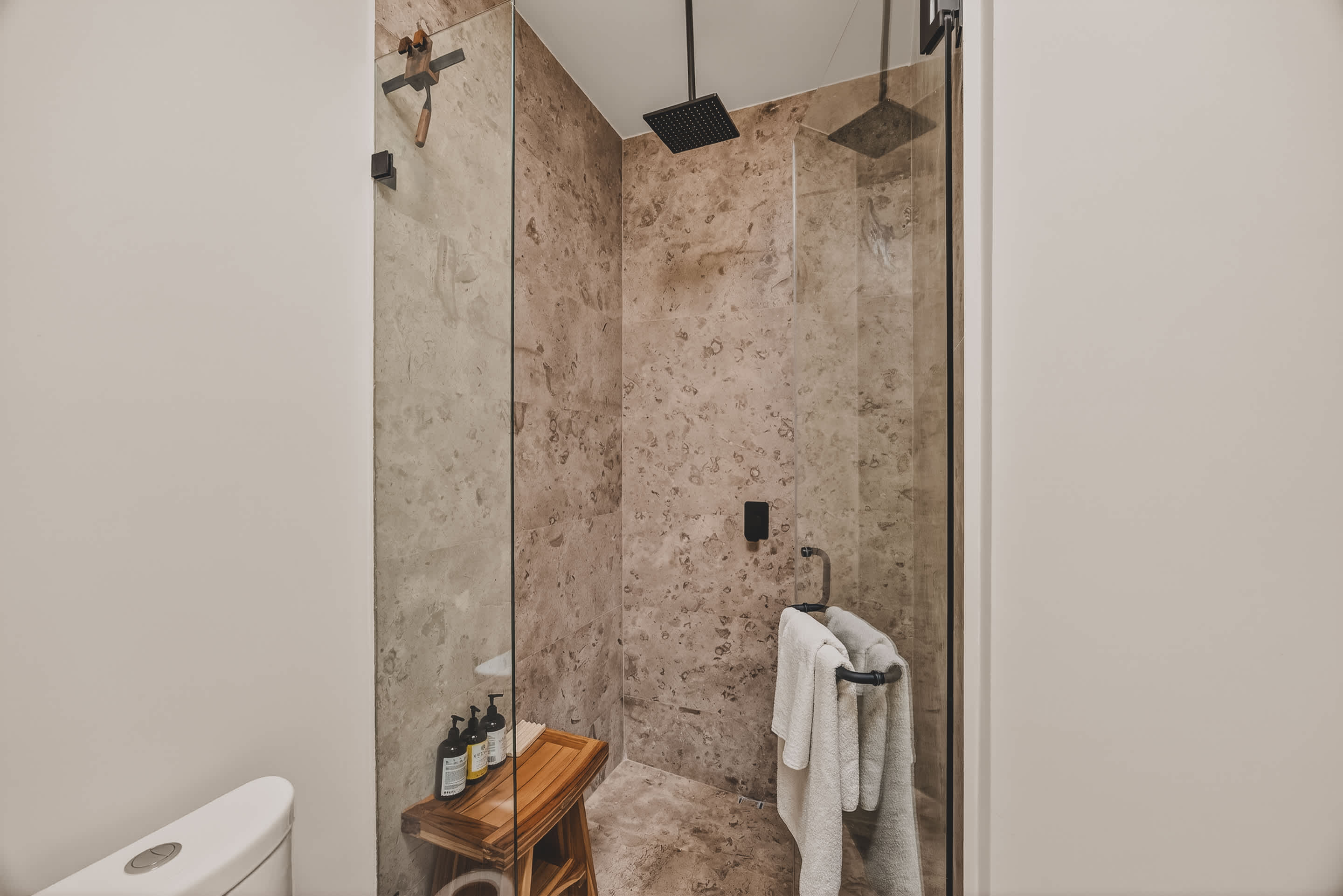 Spacious walk-in shower with light-colored stone-patterned tiles, a black rain showerhead, and a glass enclosure, featuring a small wooden stool inside.